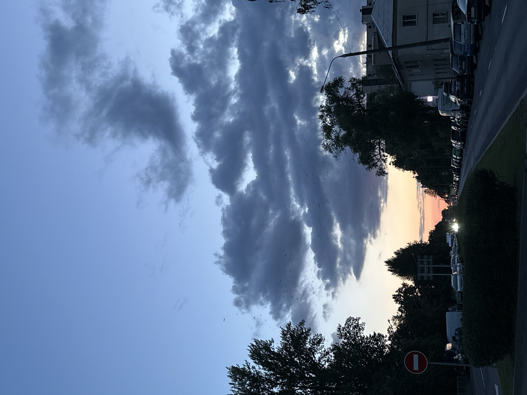 The image depicts a street scene in Minsk, Belarus, at dusk. The sky is a deep blue with dramatic, dark clouds that are beginning to break apart. A faint orange glow emanates from the horizon, indicating the setting sun. A large flock of birds is visible in the sky, dispersed among the clouds.
On the left side of the road, there are trees and a red and white circular "no entry" sign. A line of cars is parked on the right side of the road, and a traffic light is visible further down. Buildings are present on the right side of the street, some with visible windows. The road itself is paved, with visible lane markings. The overall atmosphere is calm, with a sense of the day coming to a close.