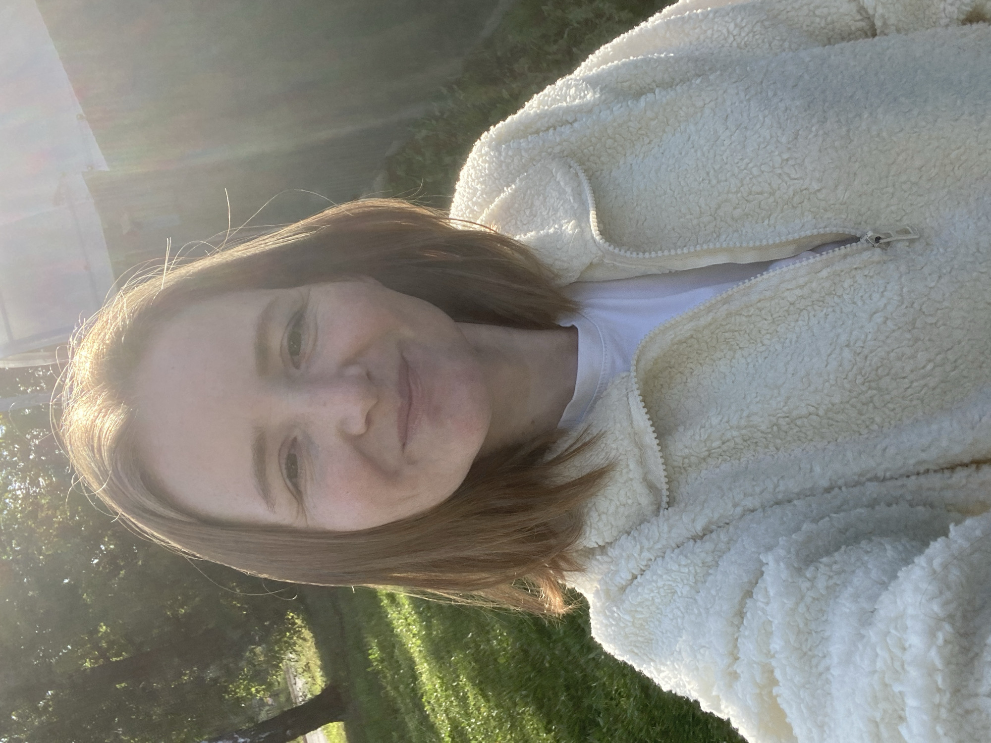 An outdoor, close-up, selfie-style photograph shows a woman with light brown, shoulder-length hair and fair skin. She is smiling and wearing a cream-colored, fuzzy jacket over a white t-shirt. The sun is shining brightly, creating a lens flare effect and casting shadows on the green grass in the background. There is a blurred fence and trees visible in the background, suggesting a residential area. The overall lighting suggests it is daytime, possibly late afternoon due to the low angle of the sun and the warmth of the light. The image is centered on the woman's face and upper body. The location is identified as Chaikovsky, Russia.