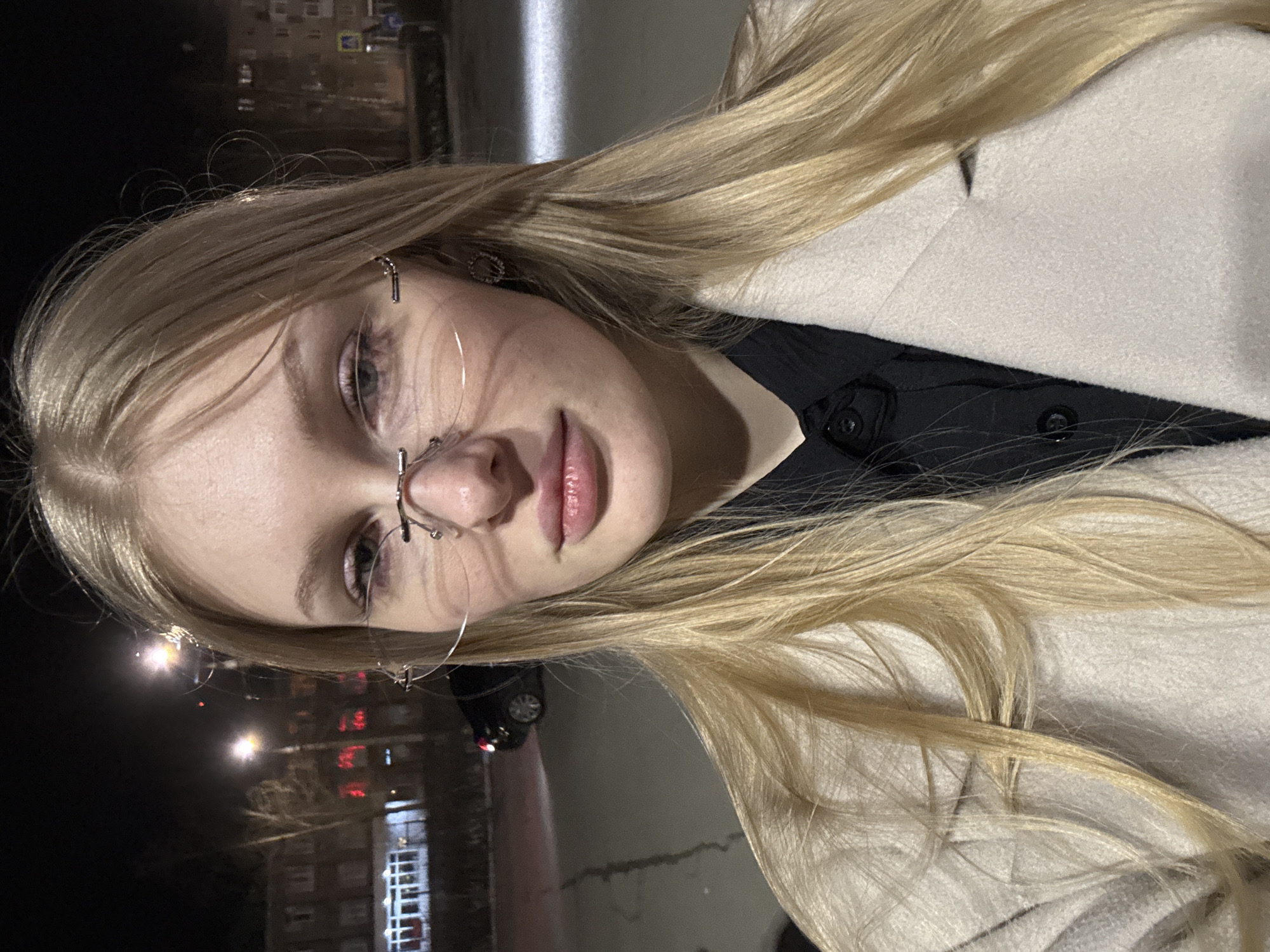 A young woman with blonde hair and silver-rimmed glasses is posing for a close-up photograph outdoors at night. She is wearing a light-colored coat over a dark shirt or dress. The background shows a street scene with multi-story buildings, streetlights, and some snow or ice on the ground. The overall lighting is low, suggesting it is after dark. Based on architectural style and ambient details such as license plates, the location appears to be in Perm, Russia.