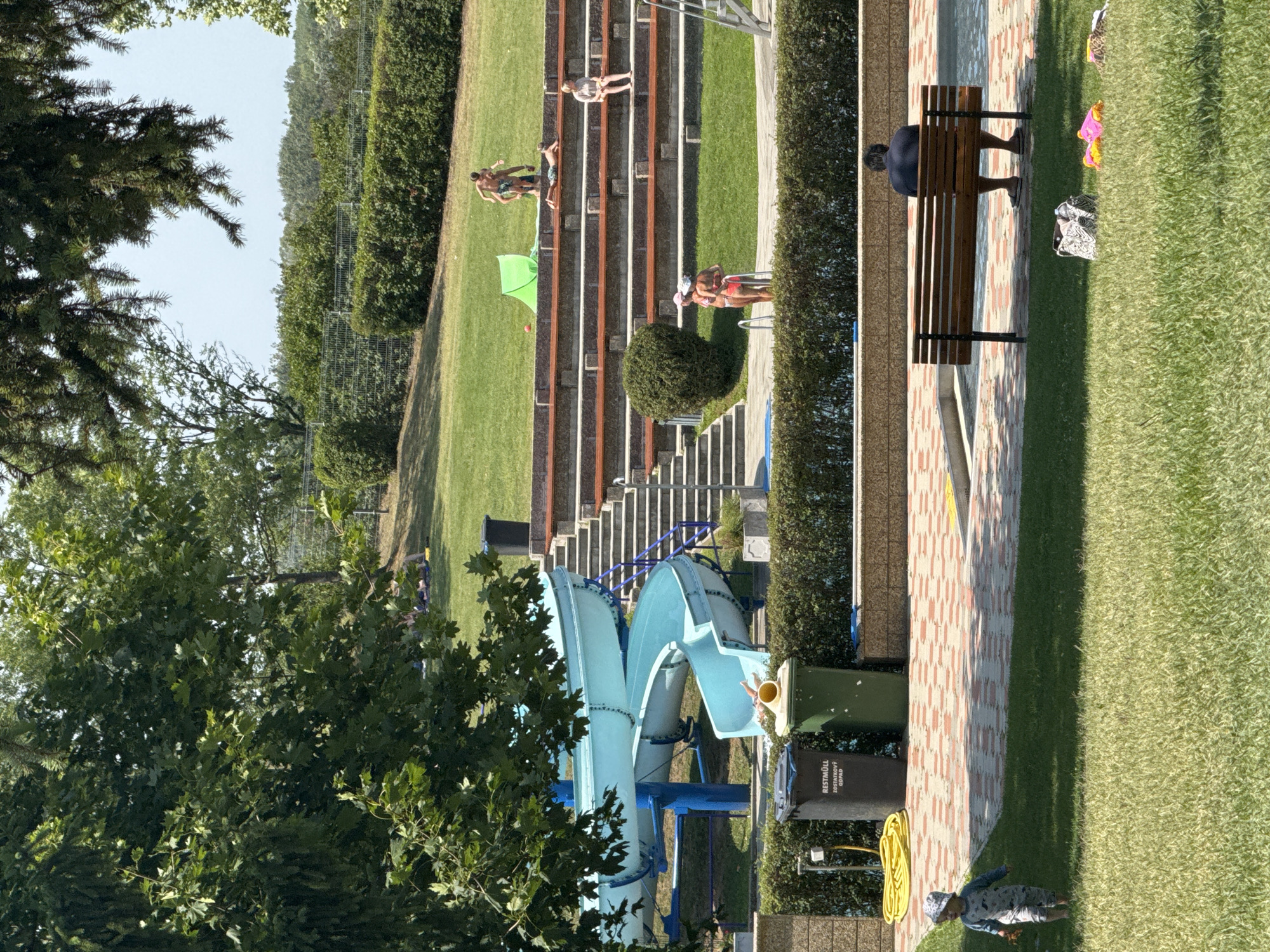 The image depicts an outdoor swimming pool complex in Hainburg an der Donau, Austria, on a sunny day. In the foreground, a young child wearing a patterned onesie and a sun hat walks across a grassy area. To the left, two large trash bins are visible, one labeled "RESTMÜLL ZOSTKOVY ODPAD". Behind the bins, a blue water slide curves downwards, with a person's legs visible at the bottom, apparently exiting the slide. A yellow coiled hose lies on the ground near the bins. In the midground, a tiered seating area with wooden benches faces the pool area. Several individuals are seated and standing on these tiers. Further back, a green tent is set up on the grass, and a few people are visible lying down or interacting. The background consists of lush green trees and bushes, with a clear blue sky above. A man sits alone on a wooden bench in the foreground, facing away from the camera. Several items, including a patterned bag and brightly colored children's toys, are scattered on the grass near the bench.