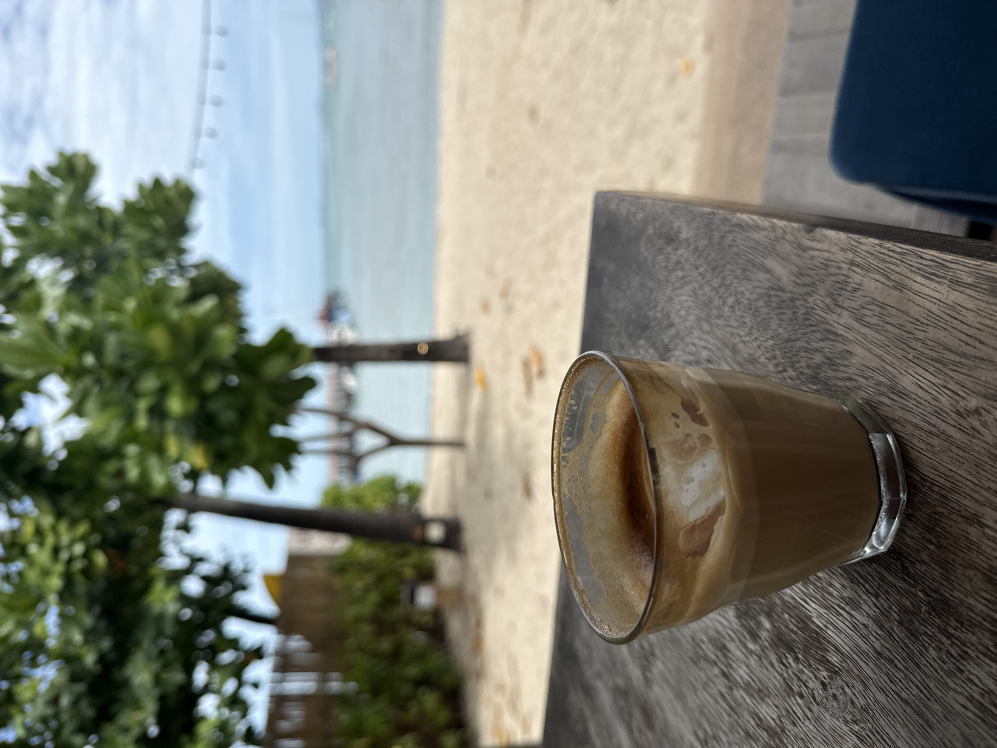 A close-up shot focuses on a glass of coffee with foam, resting on a dark, weathered wooden table. The table's surface displays a prominent wood grain texture. In the background, out of focus, a sandy beach meets a calm body of water. Lush green trees with some branches extending into the frame are visible to the left, along with a hint of a wooden structure. Further in the distance, there are faint outlines of what appear to be piers or docks and possibly some boats. The sky above is a pale blue with scattered clouds. The scene evokes a relaxed atmosphere, likely a cafe or seating area situated by the coast in a tropical location such as Koh Samui, Thailand. There are no people or animals visible in the frame.