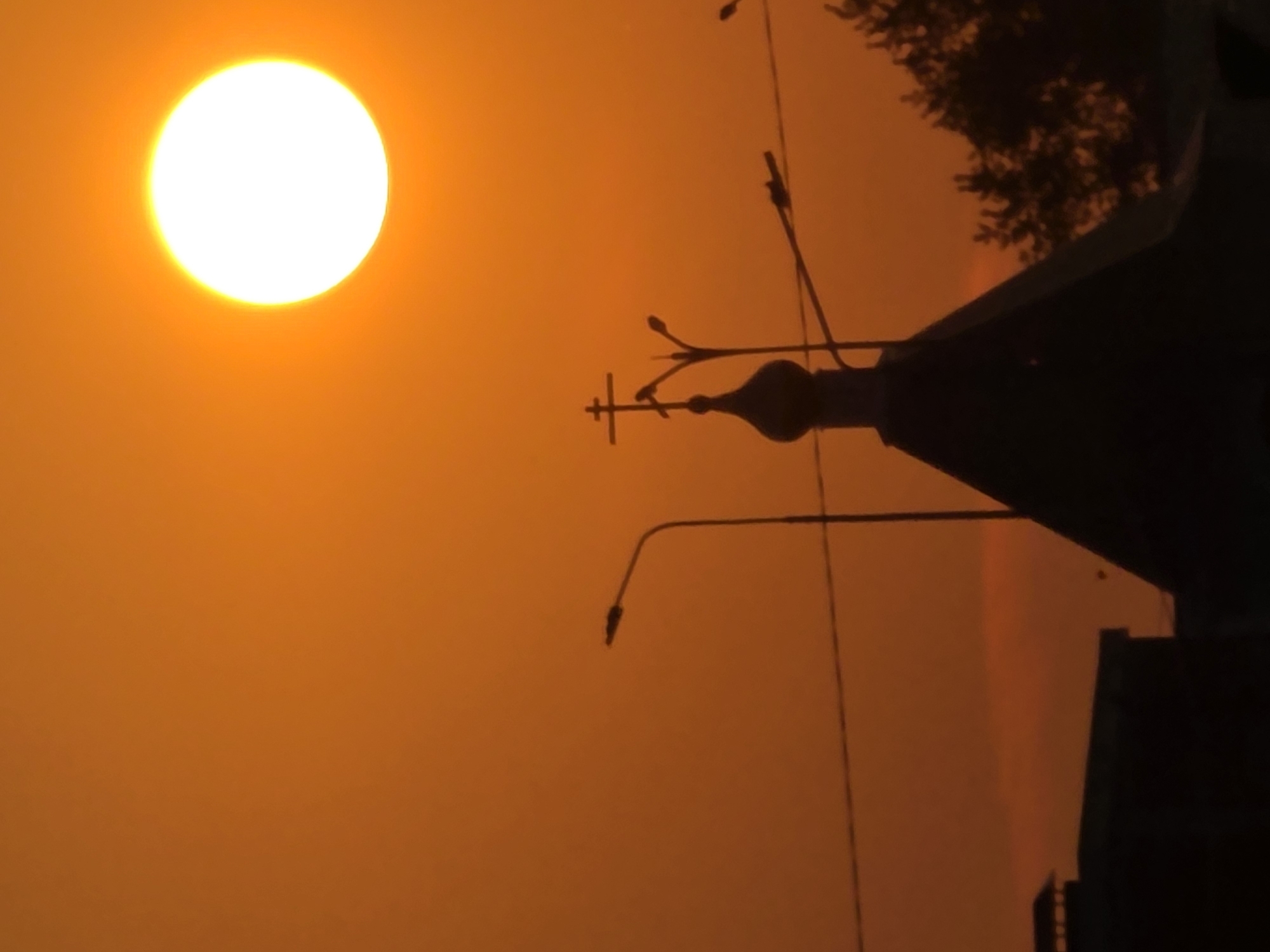 An image shows a bright, round sun setting against an orange sky. In the foreground, silhouetted against the sky, is the dark outline of a church dome with a cross on top. Two lampposts are also visible, one to the left and one to the right of the church dome. There are also some power lines stretching across the frame. The bottom of the image shows the dark silhouettes of buildings and trees, suggesting an urban or suburban setting. This scene is likely taken in Chaikovsky, Russia, given the presence of traditional Russian Orthodox architecture.