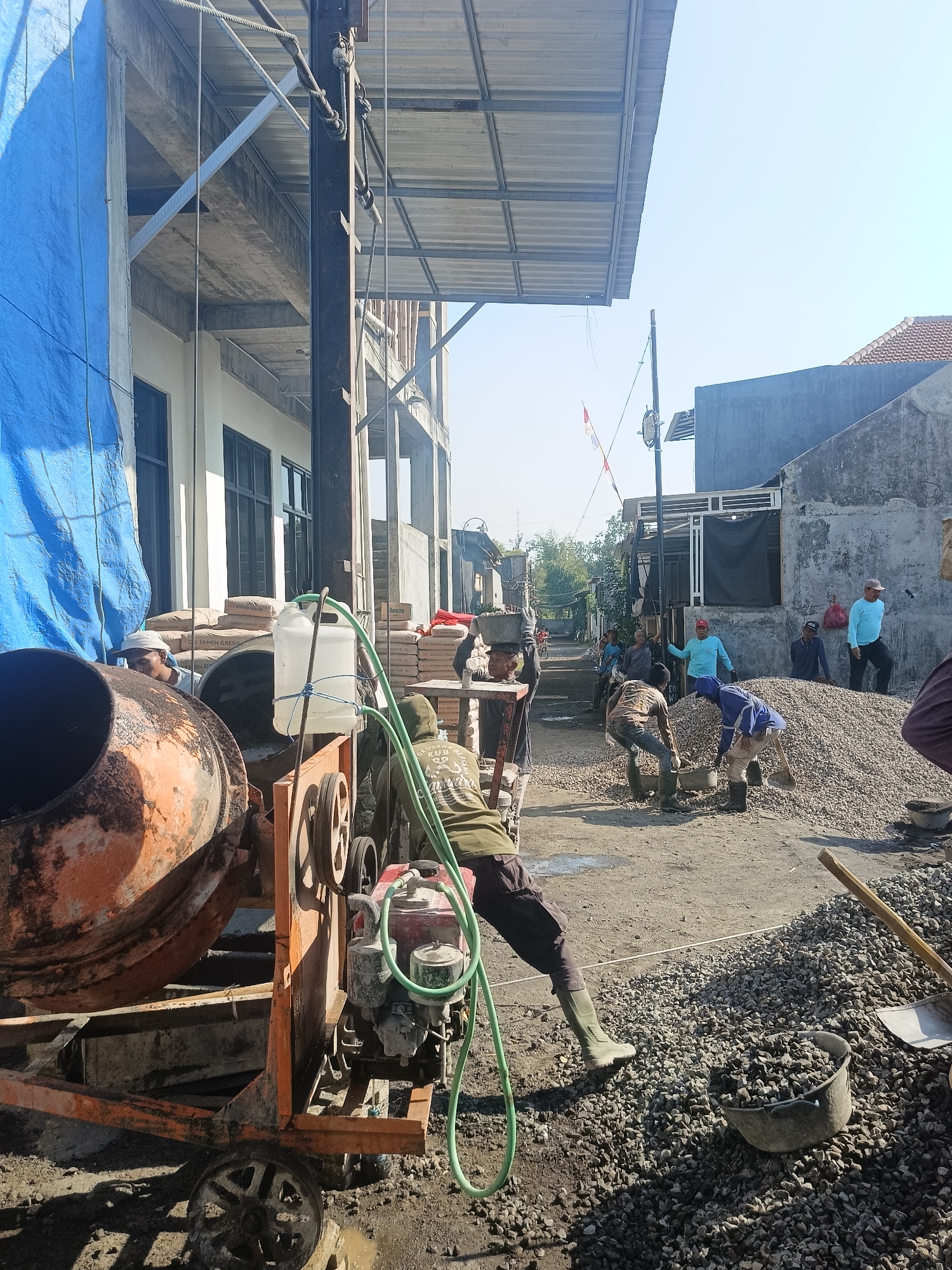 A construction site in Gedangan, Indonesia, shows a cement mixer with an orange drum and a red engine, positioned in the foreground. A white plastic water container is attached to the mixer. Behind it, several workers are engaged in construction activities. One worker, wearing a green hoodie and dark pants, is near the mixer, with a green hose connected to it. Further back, a large pile of gravel is visible. Several men in light blue shirts and one in a darker shirt are standing near the gravel. In the background, a building with a blue tarp covering part of its facade is under construction. Adjacent to this building are residential houses. The sky is clear and blue, suggesting a sunny day. A small Indonesian flag is visible on a pole in the distance.