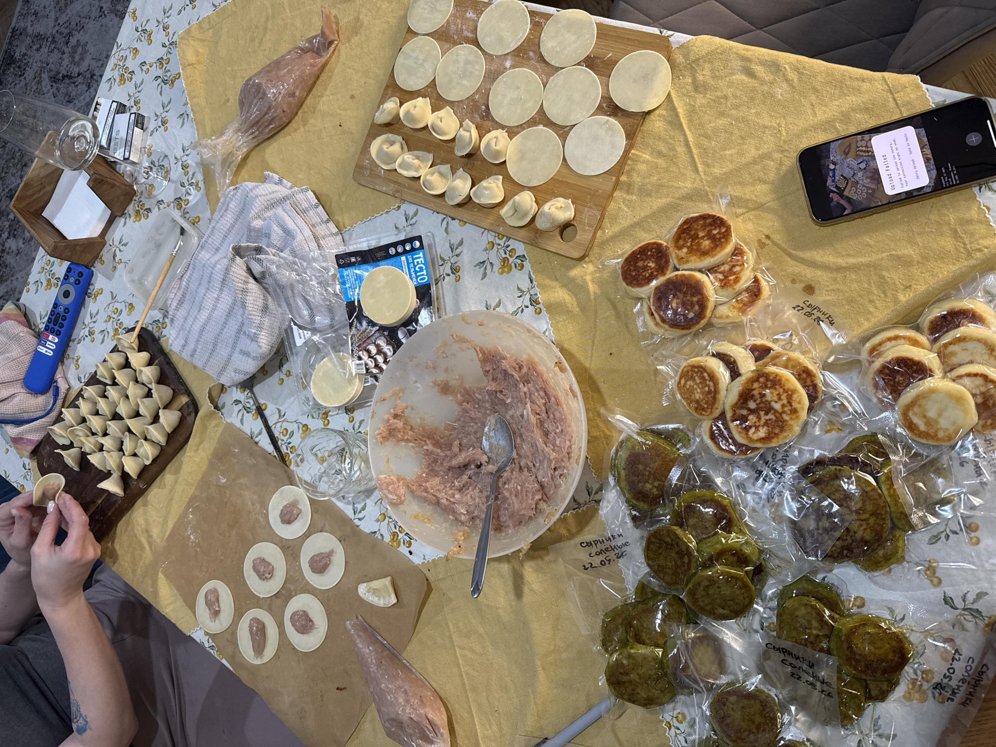 The image shows a tabletop scene with a person making dumplings ("pelmeni"). The table is covered with a yellowish tablecloth with a floral pattern. Several items are laid out on the table including: dumplings in various stages of preparation, minced meat in a large bowl, dumpling dough circles on a cutting board and parchment paper, a package of dumpling dough, a pastry bag, a wooden box, a TV remote, glasses, and a cell phone with an "Upload Failed" message. Some cooked pancakes are also visible in plastic bags. The OCR on the plastic bags indicates "Syrniki" and the date "22.05.26". Based on the OCR, the Cyrillic writing, and the items, it is likely a domestic setting in Russia or a Russian-speaking region. The city is not visible, so it is impossible to know if it's Krasnoyarsk. A person is shown making the dumplings.