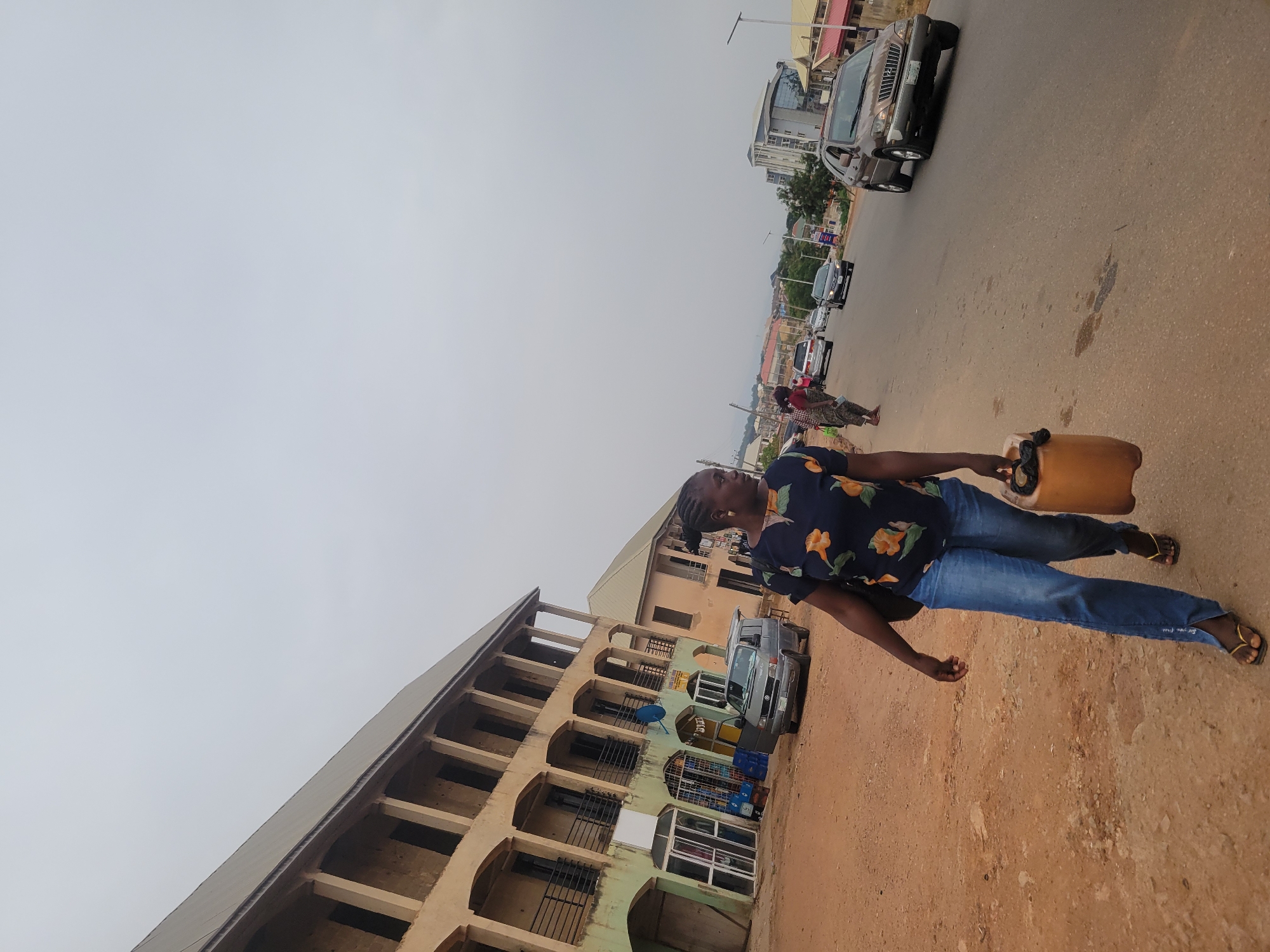 The image, rotated counter-clockwise, captures a daytime street scene in Jos, Nigeria, under an overcast sky. In the foreground, a young person, possibly female, with braided hair, walks along a dusty road surface. They are dressed in a dark blue shirt with orange floral patterns, blue jeans, and yellow flip-flops, carrying a yellow jerrycan in their right hand. Faint white text is visible near the ankle on the back of their jeans, though unreadable.
The street is paved, with visible tire marks and patches of dirt. Several vehicles are present; a dark-colored SUV is seen driving away in the mid-ground, and other cars are further down the street. Multi-story buildings line both sides of the road. On the left, a light-colored, somewhat weathered building features multiple arched windows and balconies, with security bars on the lower levels and a blue satellite dish mounted on its facade. A light-colored van is parked near this building, and goods, possibly stacked crates, are visible in front of a ground-floor shop. Other buildings, some appearing more modern, extend into the distance along the street. The general activity depicts a typical street environment with pedestrian movement and vehicle traffic.