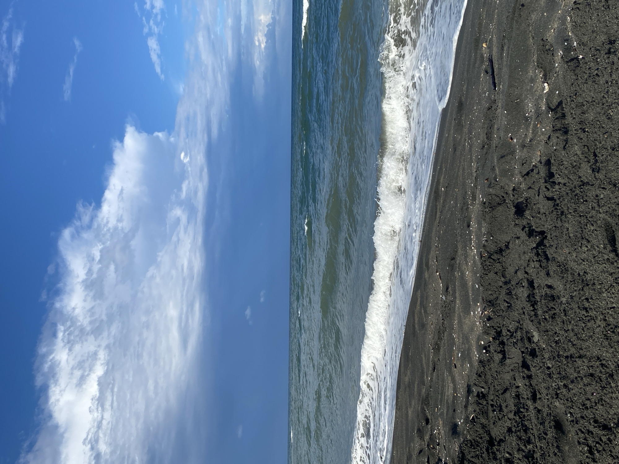 The image depicts a view of the Black Sea coast in Ureki, Georgia. The foreground is a dark, sandy beach with some small shells and debris scattered on the surface. The sand appears wet in places, indicating the proximity of the water. In the midground, the sea is a deep blue-green with moderate waves breaking on the shore, creating white foam and surf. The horizon line is clearly visible, separating the sea from the sky. The sky above is a mix of bright blue and dramatic dark clouds, suggesting a partly cloudy or potentially stormy weather condition. The overall scene is a natural landscape of a beach and the sea under a dynamic sky. There are no people or man-made structures visible in the frame.