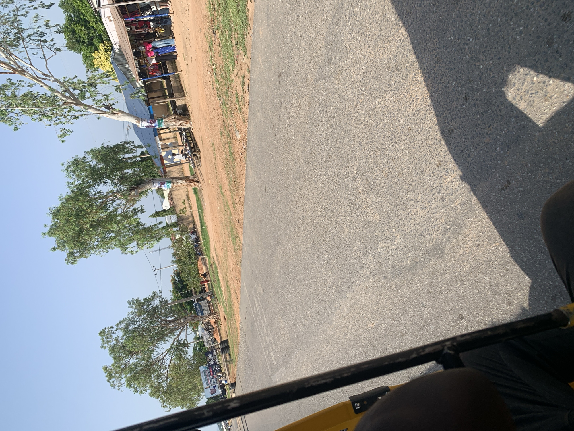 An outdoor, eye-level, full shot shows a dirt road with an asphalt lane running alongside it. On the right side of the road, there are market stalls with a variety of colorful clothing displayed. Several people are browsing and shopping in the market. Trees with thin trunks and sparse foliage are scattered throughout the scene. In the background, there are buildings, more trees, and a clear blue sky. The foreground shows the interior of a yellow vehicle, with a dark-colored bar extending from the bottom of the frame upwards to the left. The shadow of the vehicle is cast on the asphalt road. The scene appears to be in a market area within a town or city.