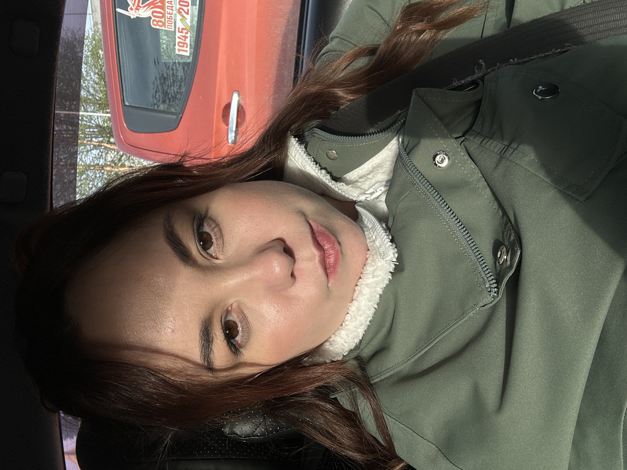 A woman is positioned inside a car. Her gaze is directed towards the camera. She is wearing a green jacket with a white fluffy collar. Part of the window is visible. Outside the window, there's an orange car with a sticker that says "80 years" and the dates "1945-2025" written on it. Trees are visible in the background, indicating the presence of nature in the scene. The location in the city is unknown.