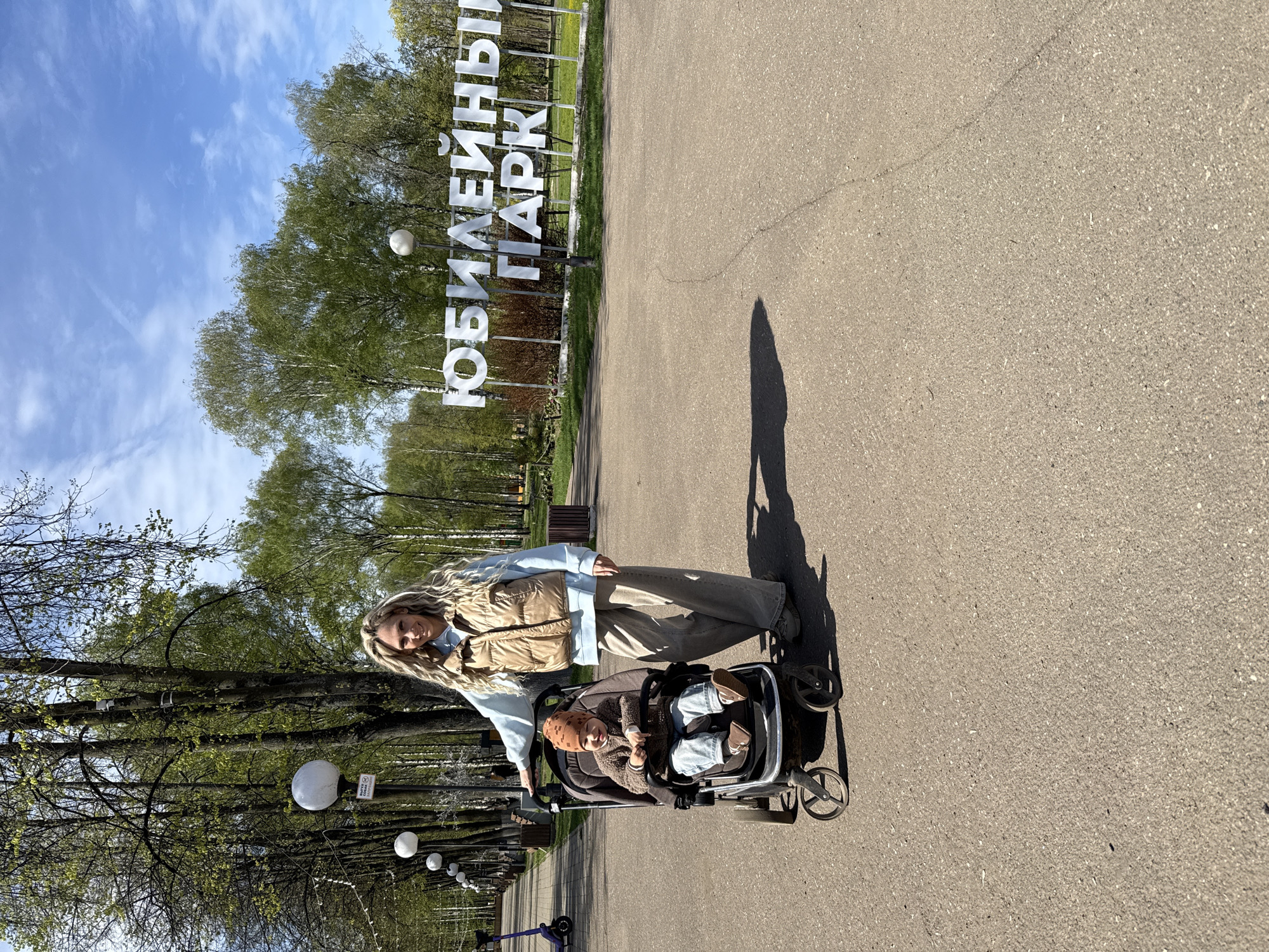 A woman is standing on a paved surface next to a baby stroller with a child in it. In the background, there is a park entrance sign that reads "ЮБИЛЕЙНЫЙ ПАРК," which translates to "Jubilee Park." The sign is white and in large letters. There are trees in the background, and the sky is visible with some clouds. The woman is wearing a light blue shirt, a beige vest, and khaki pants. The baby in the stroller is wearing a brown sweater and a hat. There are lampposts lining the side of the path, and some scooters are visible.
