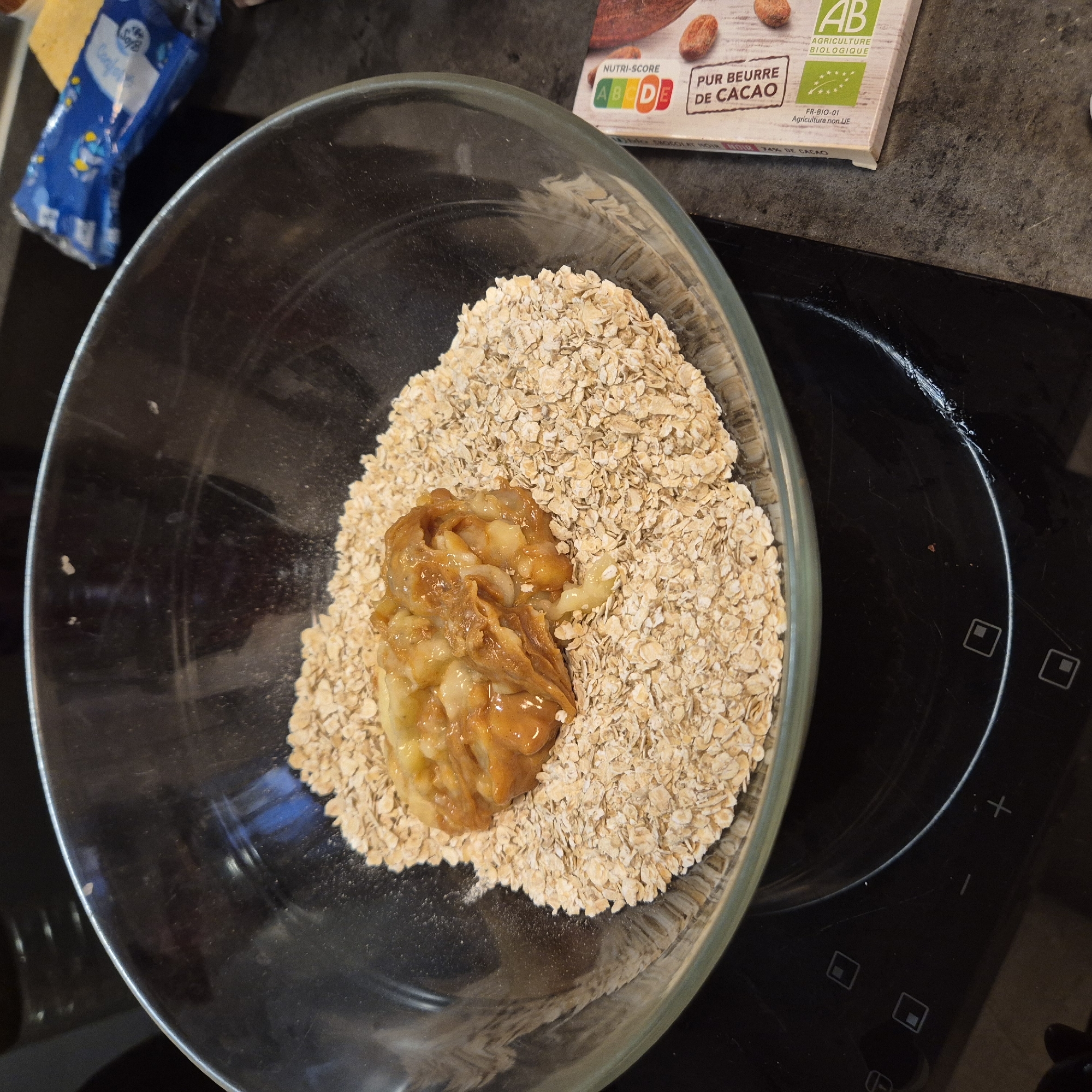A clear glass bowl sits on a black electric stovetop. The bowl contains a pile of beige rolled oats with a scoop of mashed banana and peanut butter in the center. A package of chocolate cocoa butter with a nutri-score grading sits near the bowl's edge. Partially visible nearby are a blue package of confectionary and control panel buttons on the stovetop. The presence of French product packaging suggests a location in France, likely Strasbourg.