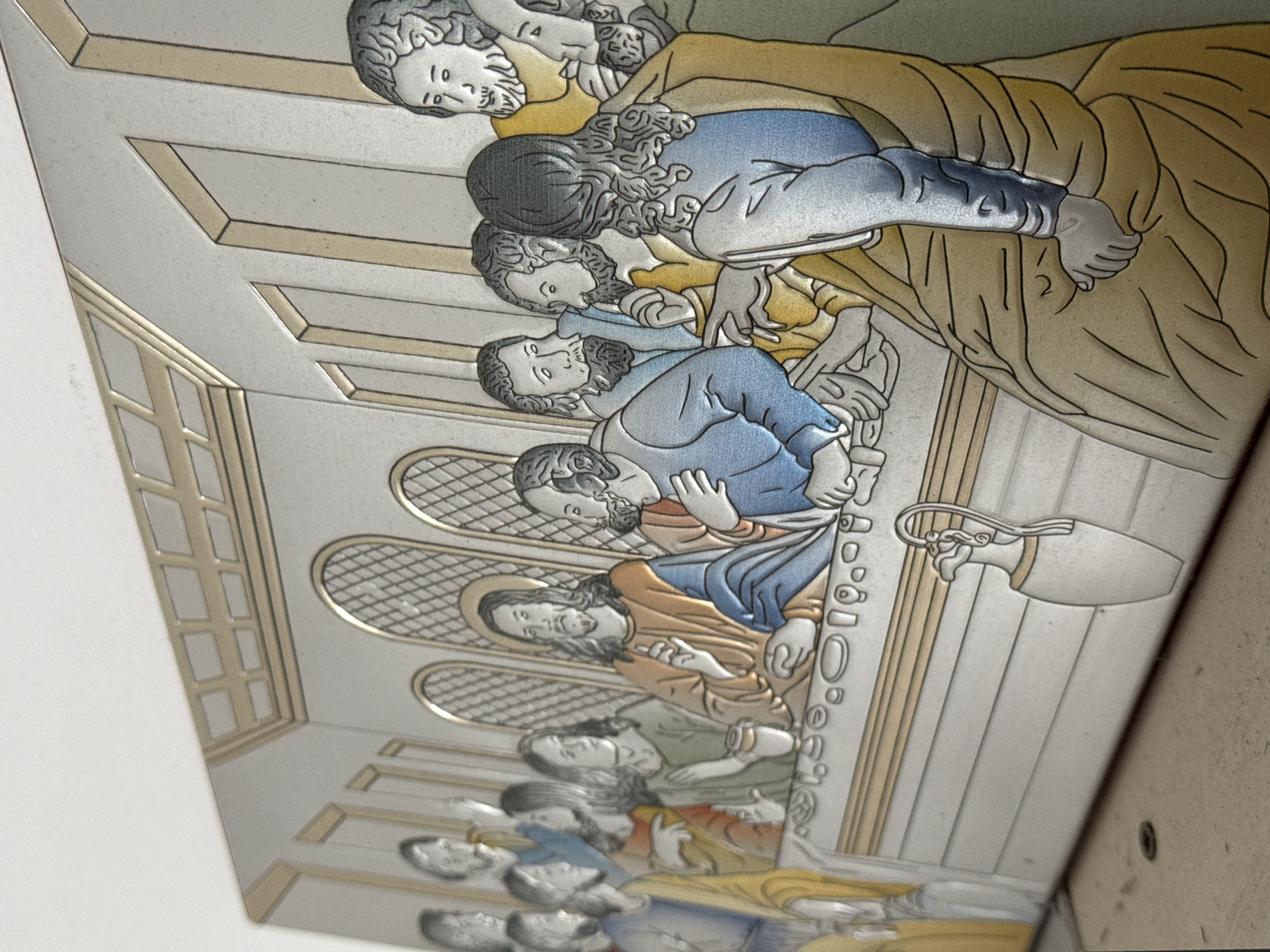 A close-up, angled shot depicts a depiction of the Last Supper, likely an artwork, possibly embossed or carved. The work is a rendition of the famous scene of Jesus and his apostles at a table. Figures are rendered in light shades of tan, yellow, blue, and gray, with distinct outlines and detailing. The scene occurs in an interior, with architectural details such as a window and ornate ceiling visible. An amphora-like vessel is visible on the side of the table. The location in Varna, Bulgaria is not apparent.