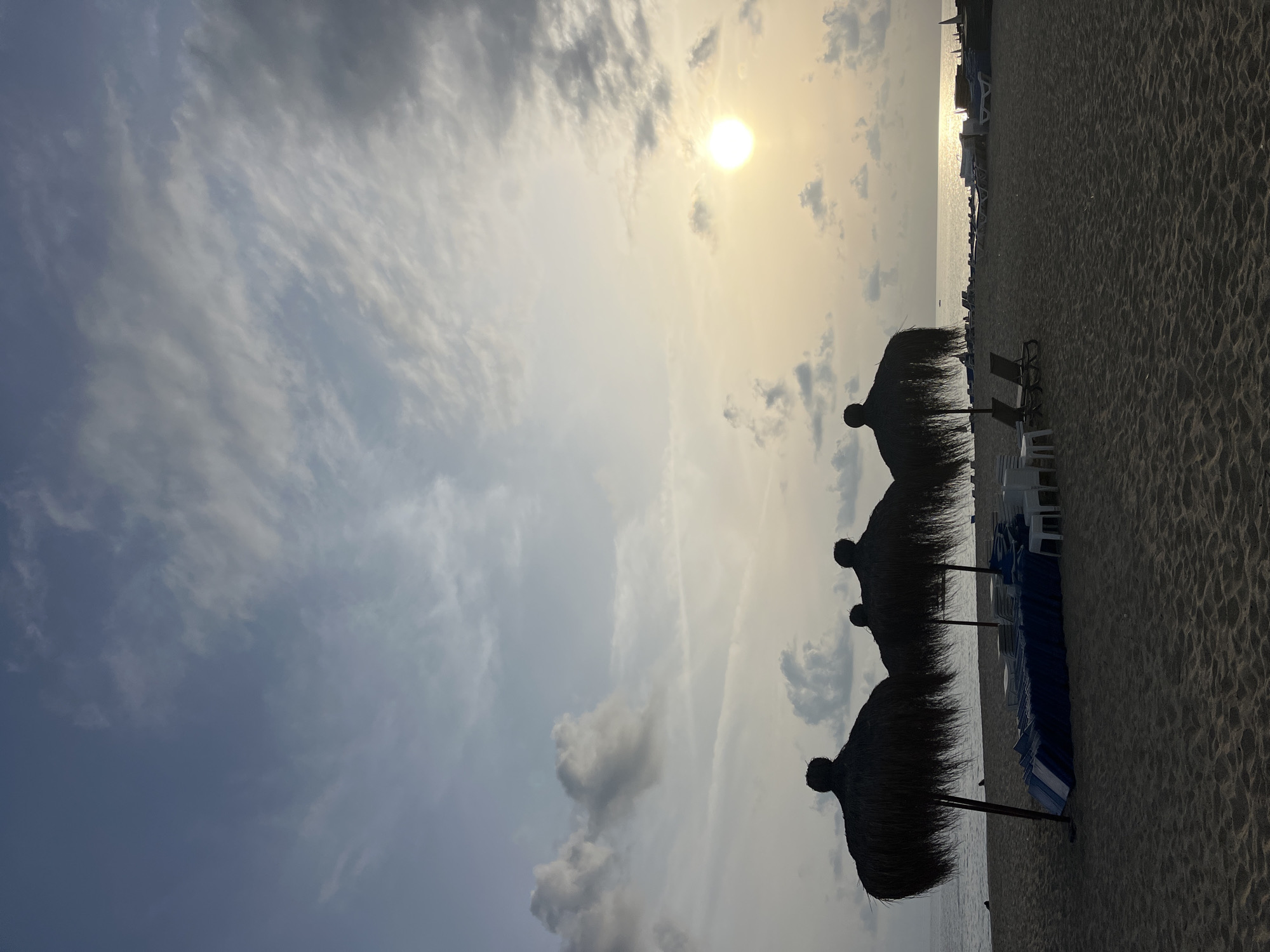 The image depicts a serene beach scene in Alanya, Turkey, likely captured during sunrise or sunset due to the low sun and warm lighting. The sky occupies a significant portion of the frame, showcasing a transition from soft blue-grey with fluffy clouds on the left to a bright, golden-yellow haze on the right. A prominent, brilliant sun is visible in the upper-right, casting a radiant glow and reflecting brightly on the calm sea.
A clear horizon line separates the sky from the tranquil sea and sandy beach. The sea stretches across the mid-ground, appearing calm and reflecting the dazzling light from the sun. In the foreground, a wide sandy beach shows subtle ripples and textures. Three large, dark, thatched beach umbrellas (palapas) are silhouetted against the bright sky and water, standing on the sand. Beyond these, numerous beach chairs and loungers, some blue and stacked, others white, are arranged along the shore.
No people are visible, lending a peaceful and quiet atmosphere to the scene. The overall impression is one of tranquility and natural beauty at a resort beach, either just beginning its day or winding down. There is no visible text in the image.