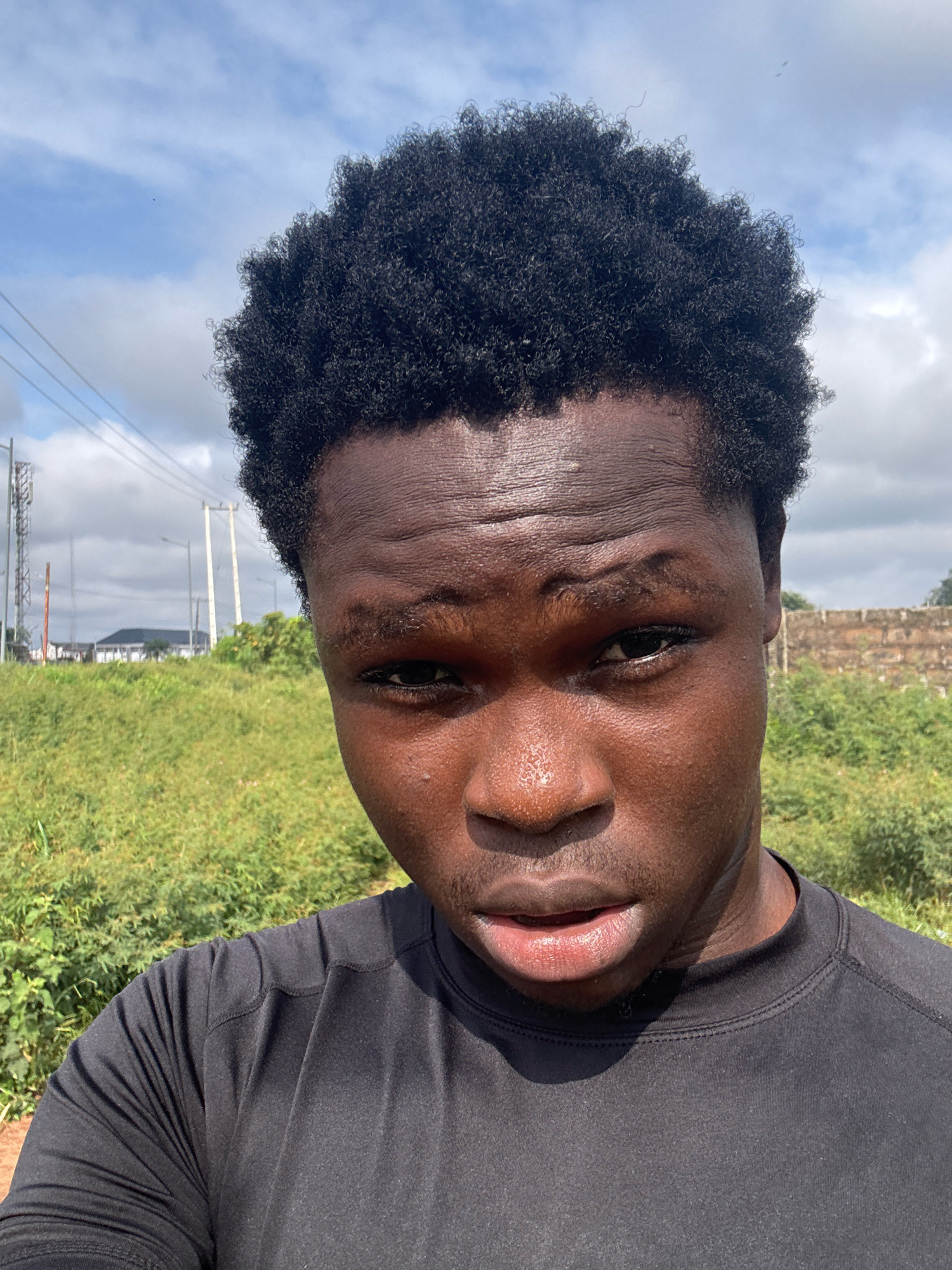 A close-up, outdoor, waist-up portrait photograph shows a dark-skinned young man with tightly curled, dark hair. He is looking directly at the camera, with sweat visible on his forehead and nose. He is wearing a black t-shirt. The background consists of a grassy field with some scattered greenery, and in the distance, a brick wall, telephone poles, and a building. The sky is visible above, with scattered clouds. The location is identified as Ihiagwa, Nigeria.