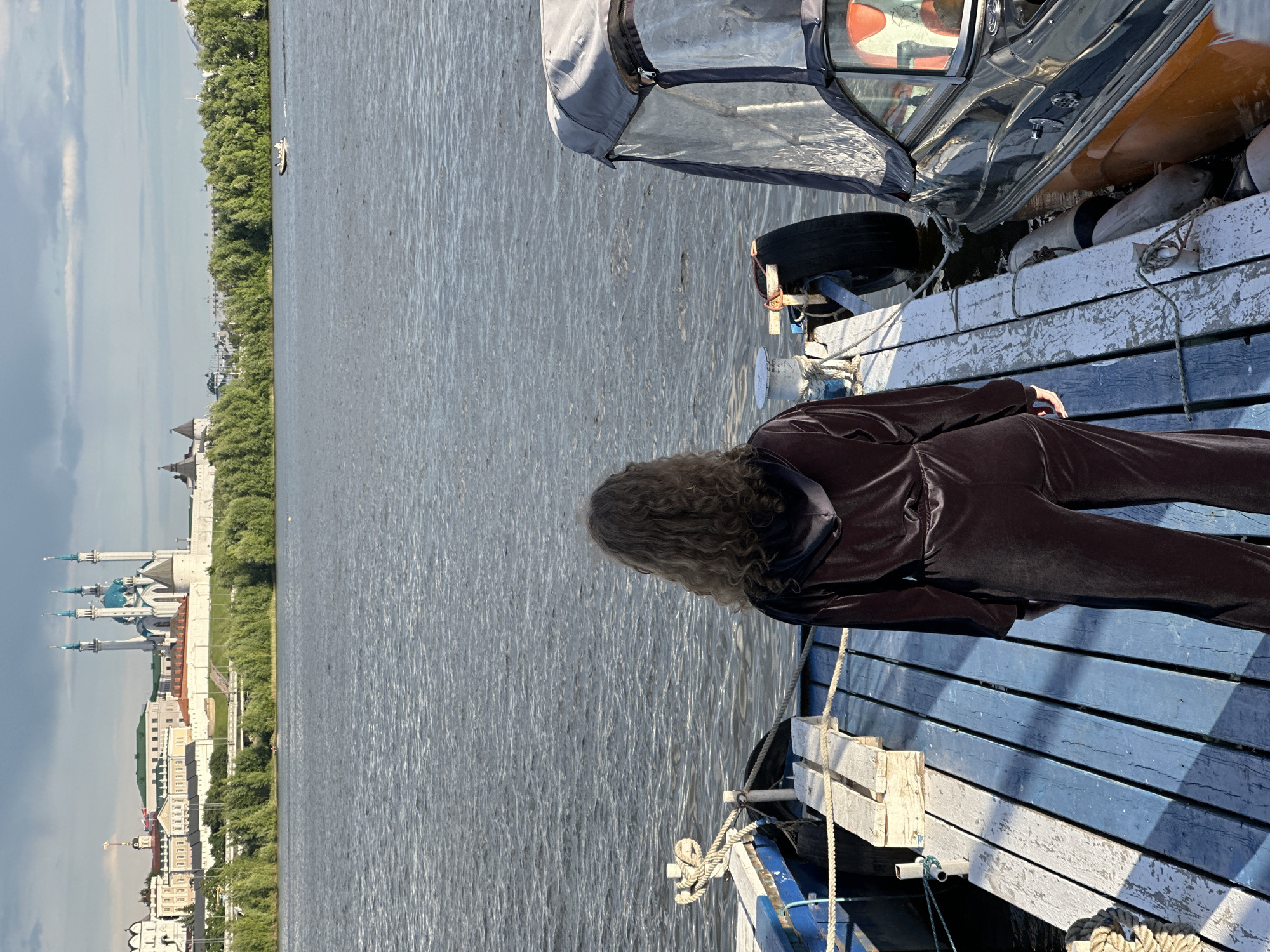 A slightly rotated medium shot captures a woman standing on a blue and white wooden dock. She is dressed in a dark brown or black velvet tracksuit, facing away from the camera towards a body of water. Her long, curly hair cascades down her back. Beside her, on the dock, sits a boat, partly covered with a gray protective cover and various buoys and marine equipment. In the distance, across the water, lies a city skyline. The skyline is marked by the distinct architecture of the Kazan Kremlin, identifiable by its white stone walls and the iconic blue-domed Kul Sharif Mosque. The city is Kazan, Russia. The weather appears fair, with a mix of sun and clouds.
