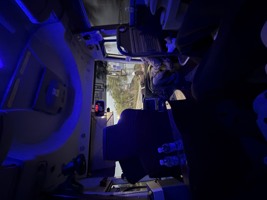 The image depicts the interior of a passenger bus illuminated by blue LED accent lighting along the ceiling and overhead compartments. The perspective is from a middle-section passenger seat, looking forward. In the front, a digital clock displays "07:53" above the windshield. A male driver is visible, partially obscured, operating the vehicle. Through the front windshield, a tree-lined road is visible on the left, suggesting motion. To the right, another passenger is seated in the front-right seat, visible from the torso down, wearing blue jeans and light-colored shoes. The external view through the right-side window shows a dark environment with distant lights, possibly over water. In the immediate foreground-left, two clear plastic water bottles rest on a surface. The environment indicates travel within Tuan Chau Ward, Vietnam.