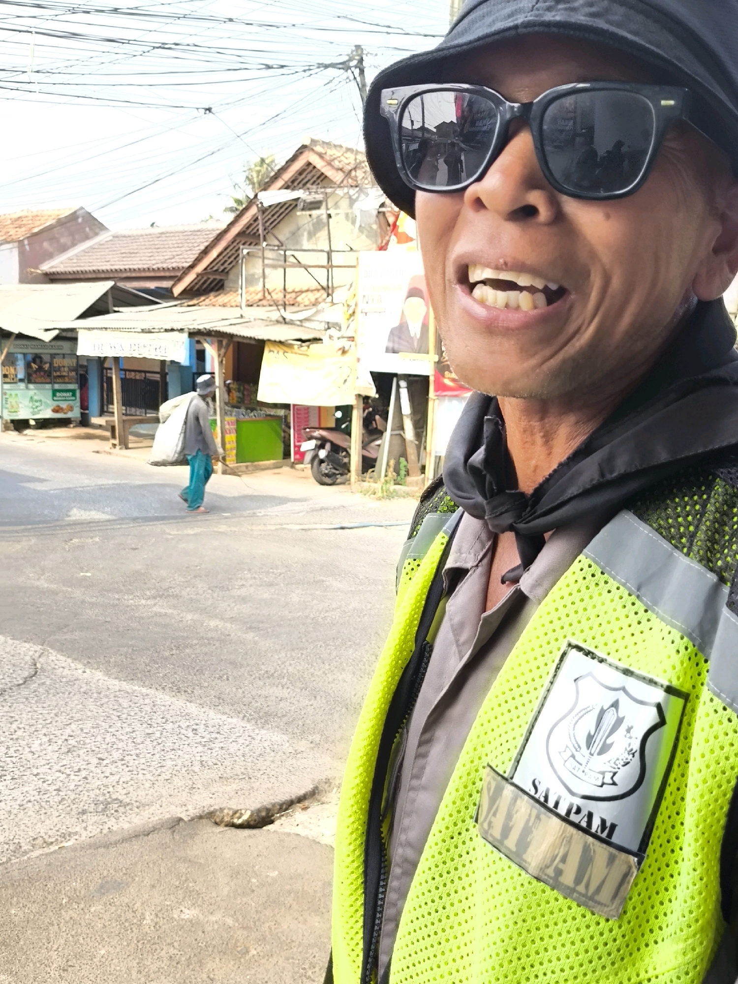 In Lubangbuaya, Indonesia, a man wearing a black bucket hat and black sunglasses smiles, revealing his teeth. He is also wearing a grey shirt and a bright yellow safety vest with reflective grey trim. The vest has a badge with a shield emblem and the text "SATPAM" above it. Below the badge is a strip of fabric with the word "SATPAM" in block letters. In the background, a street scene unfolds with several buildings, some with corrugated metal roofs. A person in a grey long-sleeved shirt and teal pants walks away from the camera, carrying a large white sack over their left shoulder. A motorbike is parked on the right side of the street. Overhead, a complex network of electrical wires crisscrosses the sky. The road surface shows signs of wear, with cracks and uneven patches.