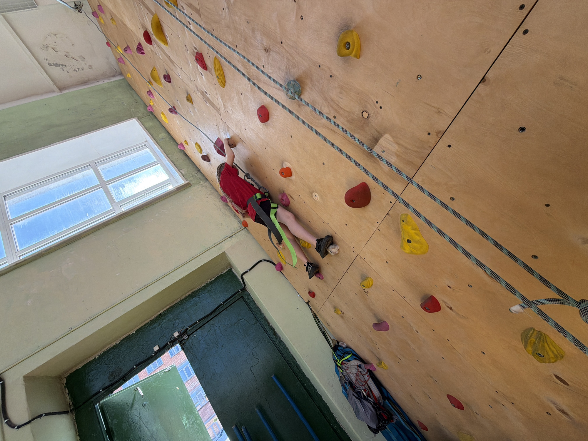 A young person is climbing an indoor climbing wall. The wall is constructed of plywood and features a variety of climbing holds in different colors and shapes. The climber is wearing a red shirt, climbing harness, and climbing shoes. They are attached to a rope that is anchored at the top of the wall.
The location appears to be an indoor gymnasium or recreational facility, with a window and door visible. The overall scene is angled. The location is likely Izhevsk, Russia.
