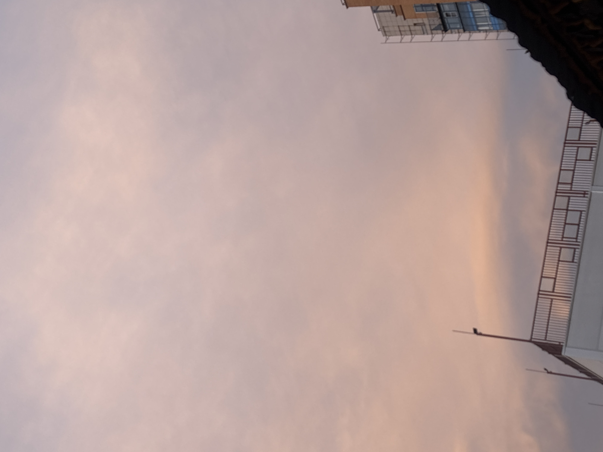 The image shows a view of the sky at what appears to be dusk or dawn, with the light having a pinkish-orange hue. In the top right corner, there is a fragment of a building, possibly an apartment complex, with windows and balconies visible. Adjacent to this, there's a structure that seems like a bridge or elevated walkway with a metal railing. Lower right has parts of unidentified buildings.
Without more context or landmarks, the location within Krasnoyarsk, Russia, cannot be definitively determined.
