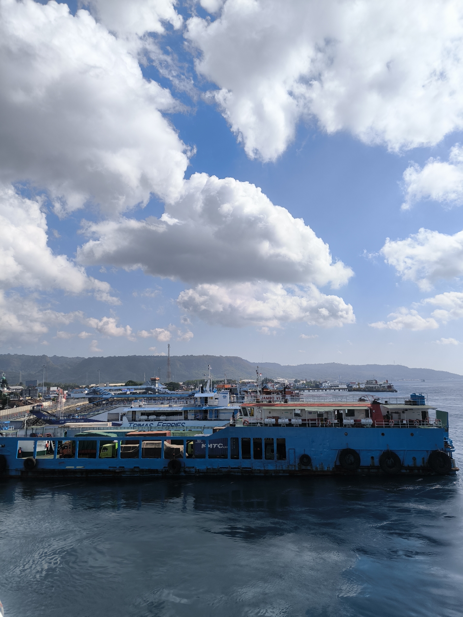 This image captures a view of the Ketapang Ferry Port in Ketapang, Indonesia, under a bright, cloudy sky. The foreground is dominated by a large, blue ferry, its hull a solid color with visible tire fenders along its side. Through its open windows, the silhouettes of trucks and cargo can be seen. The ferry's upper decks are visible, painted in white and red, with railings and windows.
In the midground, a line of other ferries and port infrastructure extends along the waterfront. These vessels also appear to be ferries, some painted white with blue accents. The background reveals a densely populated coastline with numerous buildings, some appearing to be warehouses or port facilities, and beyond them, lush green hills or mountains rise against the horizon. The sky is a vibrant blue, scattered with large, fluffy white clouds that cast shadows on the water and the landscape. The water itself is a deep blue, showing gentle ripples and reflections. The overall scene depicts a busy port environment on a clear day.