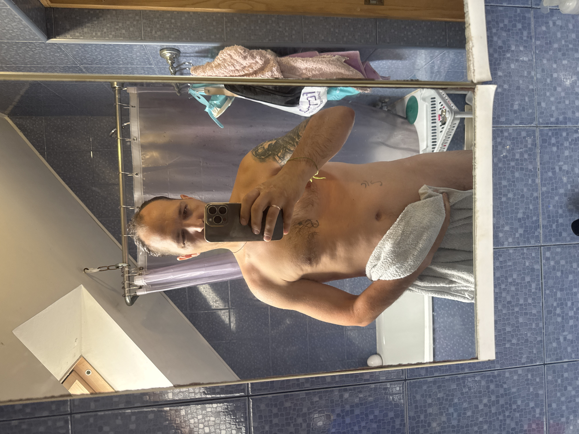 A man is taking a mirror selfie in a bathroom. He is topless and holding a towel in front of him. He has a tattoo on his left arm and a ring on his left ring finger. The bathroom has blue tiled walls and a bathtub. There is a skylight in the ceiling. The image is taken from the chest up, and the man is looking at his phone.