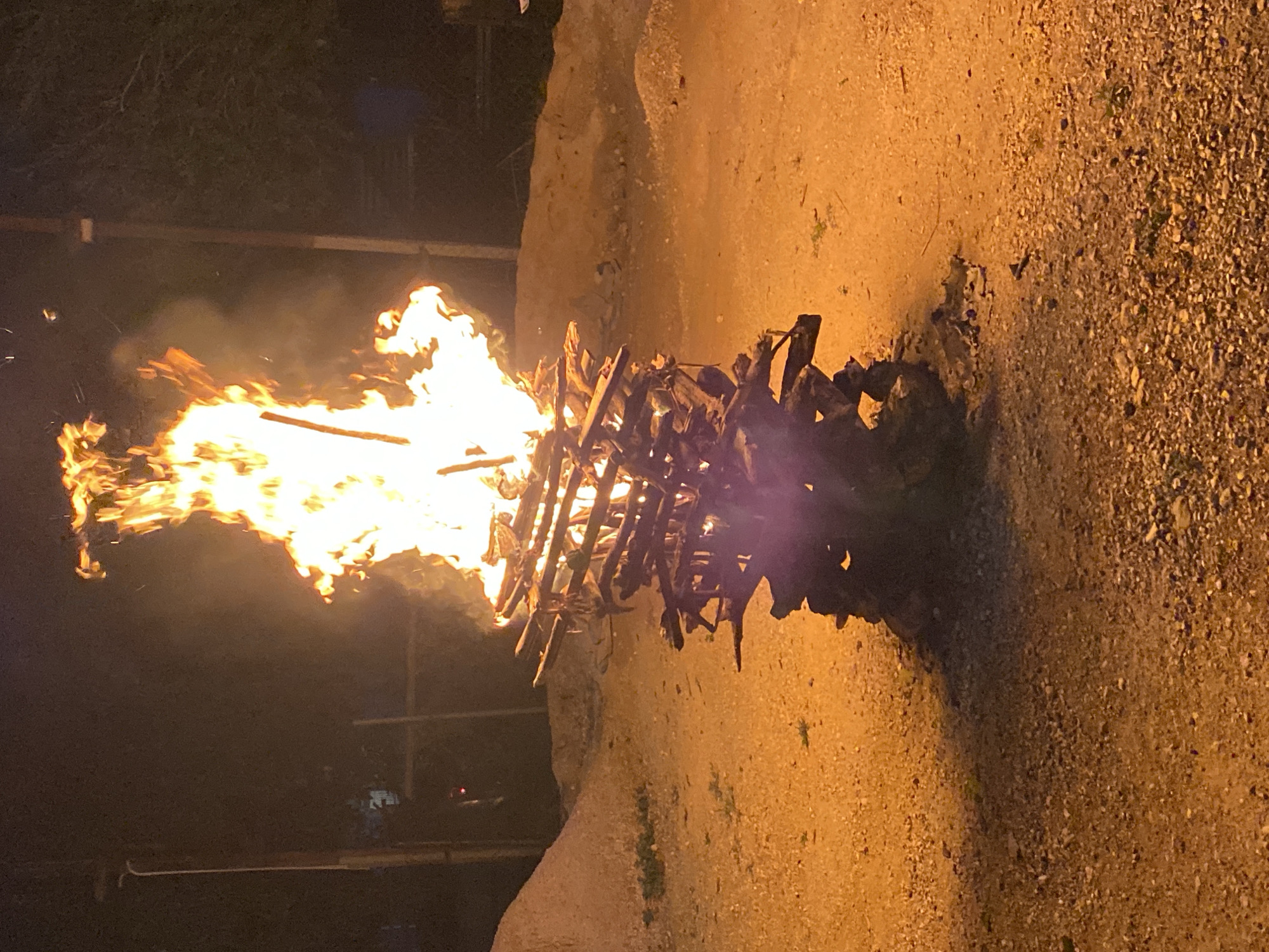 A bonfire burns brightly outdoors on an uneven, gravelly, dirt surface. The fire is built upon a stack of wooden materials. The location is Tinaquillo, Venezuela. The background is dark and contains blurry, indistinct objects. No people are visible.