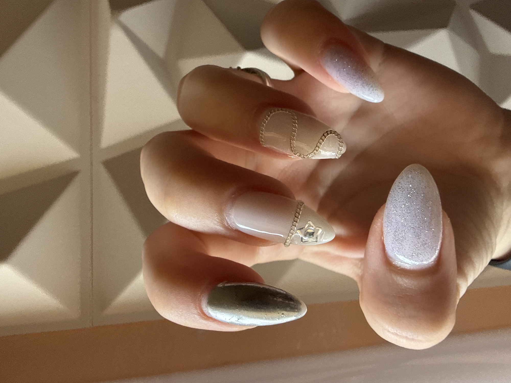 A hand with elaborately decorated fingernails is displayed. The nail on the thumb is painted in a silver metallic finish. The index finger's nail is decorated with a pale base color and a delicate gold chain around the cuticle. The middle finger's nail has a nude color with a matching gold chain outlining the base. The ring finger has a white glitter finish. The background appears to be a wall with raised geometric shapes.