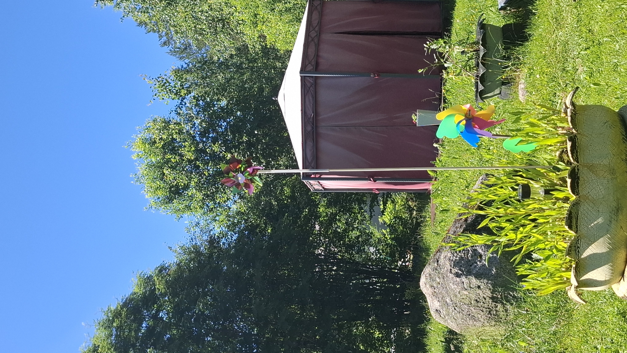 This image depicts a backyard scene on a sunny day in Novovilgovskoye Sel'skoye Poseleniye, Russia. In the foreground, a cracked, light-colored planter, possibly made from a repurposed tire, is filled with vibrant green, thin-leaved plants. A small solar-powered garden light is visible among the plants. To the left of the planter is a large, rough-textured grey boulder. A multi-colored pinwheel on a white pole stands in front of a large, hexagonal burgundy gazebo with a white roof. Behind the gazebo, dense green trees form a natural backdrop under a clear blue sky. To the right of the gazebo, another planter, also appearing to be made from stacked tires, contains a small plant with sparse leaves. The ground is covered in lush green grass, indicating a well-maintained outdoor space.
