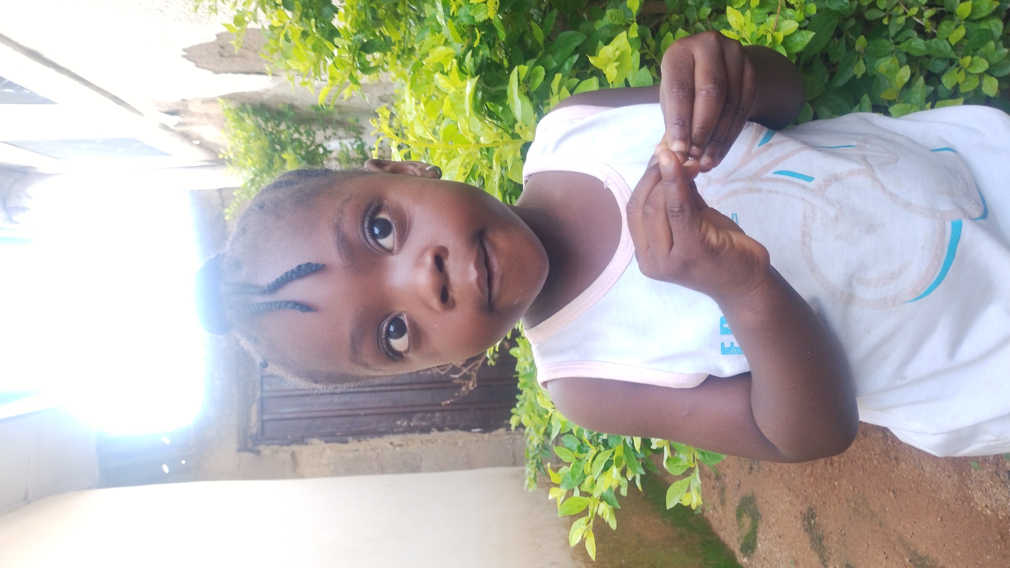 A young, dark-skinned child stands outside. They are facing forward, holding something small between their fingers. The child wears a white sleeveless top with pink trim and a blue design. They have dark eyes and braided hair. Behind the child is a green leafy shrub, a wall, and a closed window. The location appears to be in Jos, Nigeria. The ground is dirt and grass.