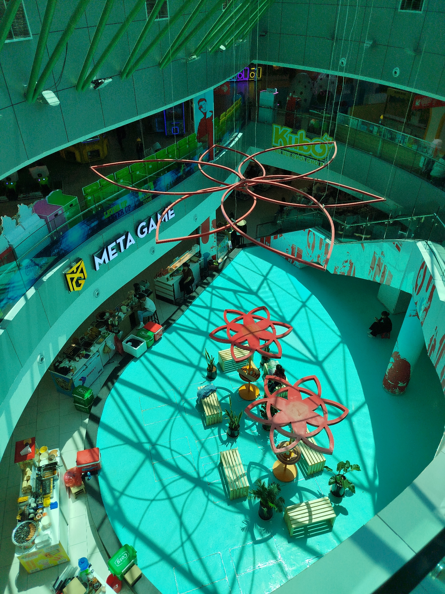 This high-angle shot depicts an indoor atrium in a shopping center in Da Lat, Vietnam, characterized by teal-colored flooring and a multi-level structure.
Several commercial establishments are visible. "META GAME" is displayed in white lettering on one side. Also present is signage for "Kubo".
The atrium features red flower-shaped decorative elements hanging overhead and as seating, with wooden benches arranged around them. Potted plants are scattered near the seating areas.
A few people are present: some stand at food stalls along the edge of the teal floor. One individual is seated against a wall.
Shadows from the overhead structure cast a geometric pattern across the floor.