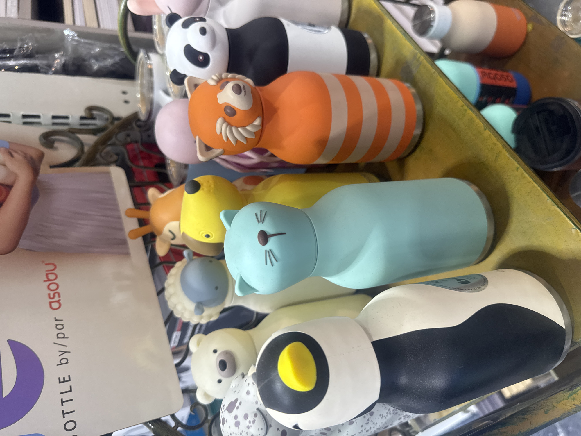 The image shows a collection of animal-shaped water bottles displayed on a shelf or tray, likely for retail. The bottles include designs of a panda, red panda, deer, cat (blue), polar bear, and an unidentified animal. The branding "BOTTLE by/par asobu" is visible on a nearby package. The setting is possibly a store in Tel-Aviv, Israel, but this is not definitively confirmed in the image itself.