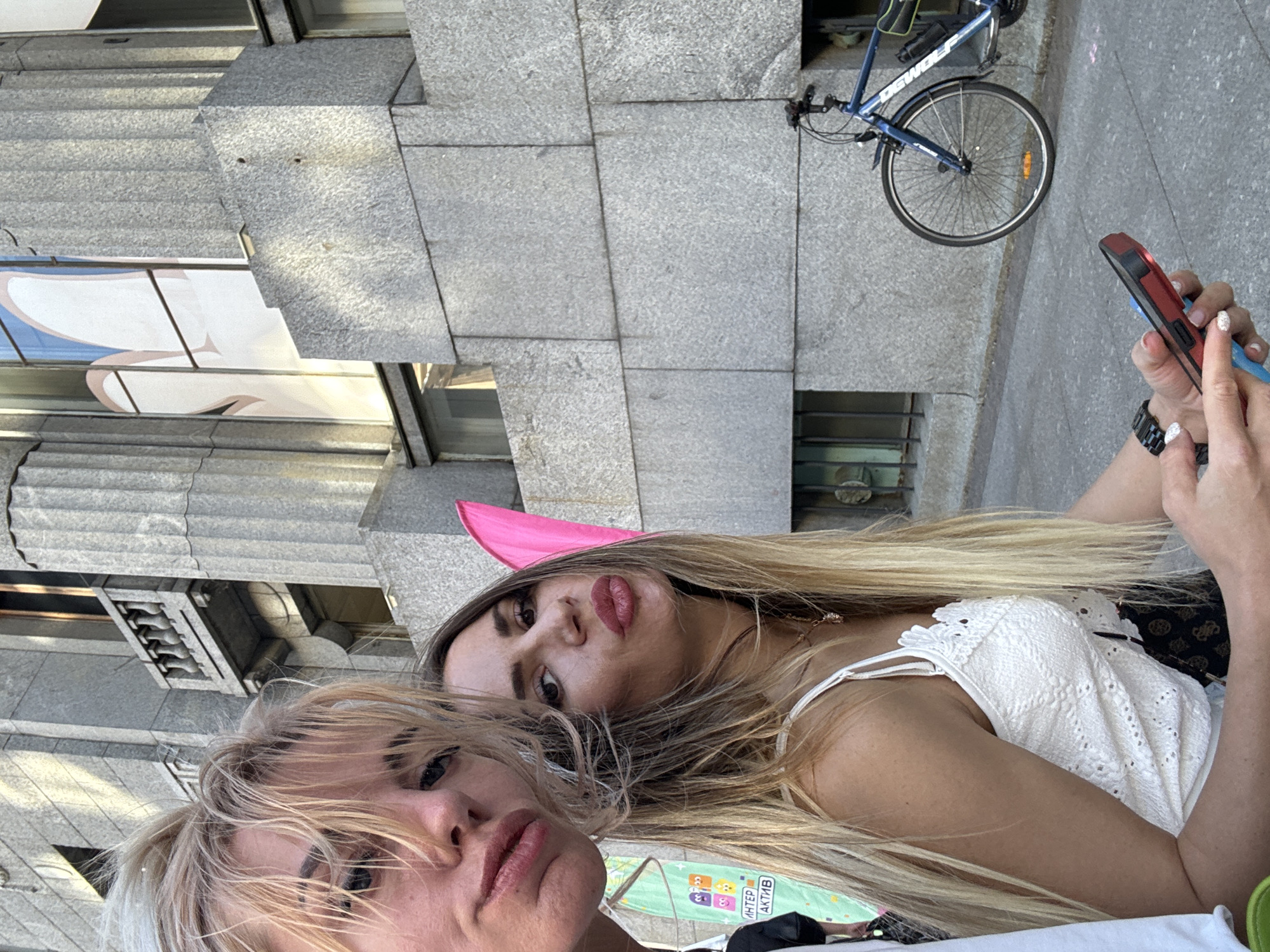 Two women are taking a selfie in front of a granite building in Saint Petersburg, Russia. The woman on the left has blonde hair and is wearing a white top. The woman on the right has long blonde hair and is wearing a white lace top. She has a pink accessory behind her head. A blue bicycle is parked on the sidewalk to the right of the women. The building appears to be an office building or a public institution, with large granite blocks and windows. The sidewalk is paved with grey stones.