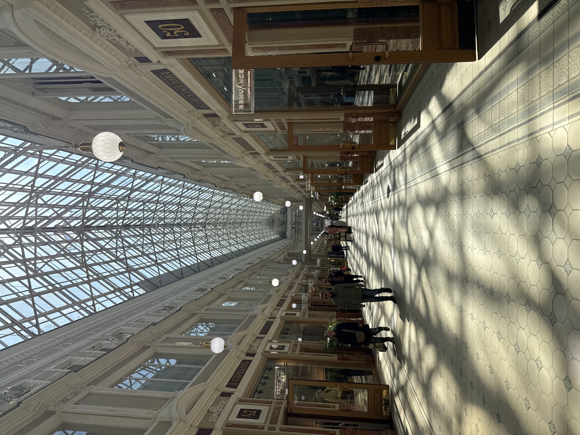 The image shows an interior view of the Passage shopping arcade in Saint Petersburg, Russia. The arcade has a high, glass-paneled arched roof that allows daylight to illuminate the tiled floor and shops lining the sides. People are walking along the corridor. The shops have display windows and signage in both Russian and English. Ornate architectural details, including moldings and framed panels, adorn the walls. Round, white hanging lamps are suspended from the ceiling.