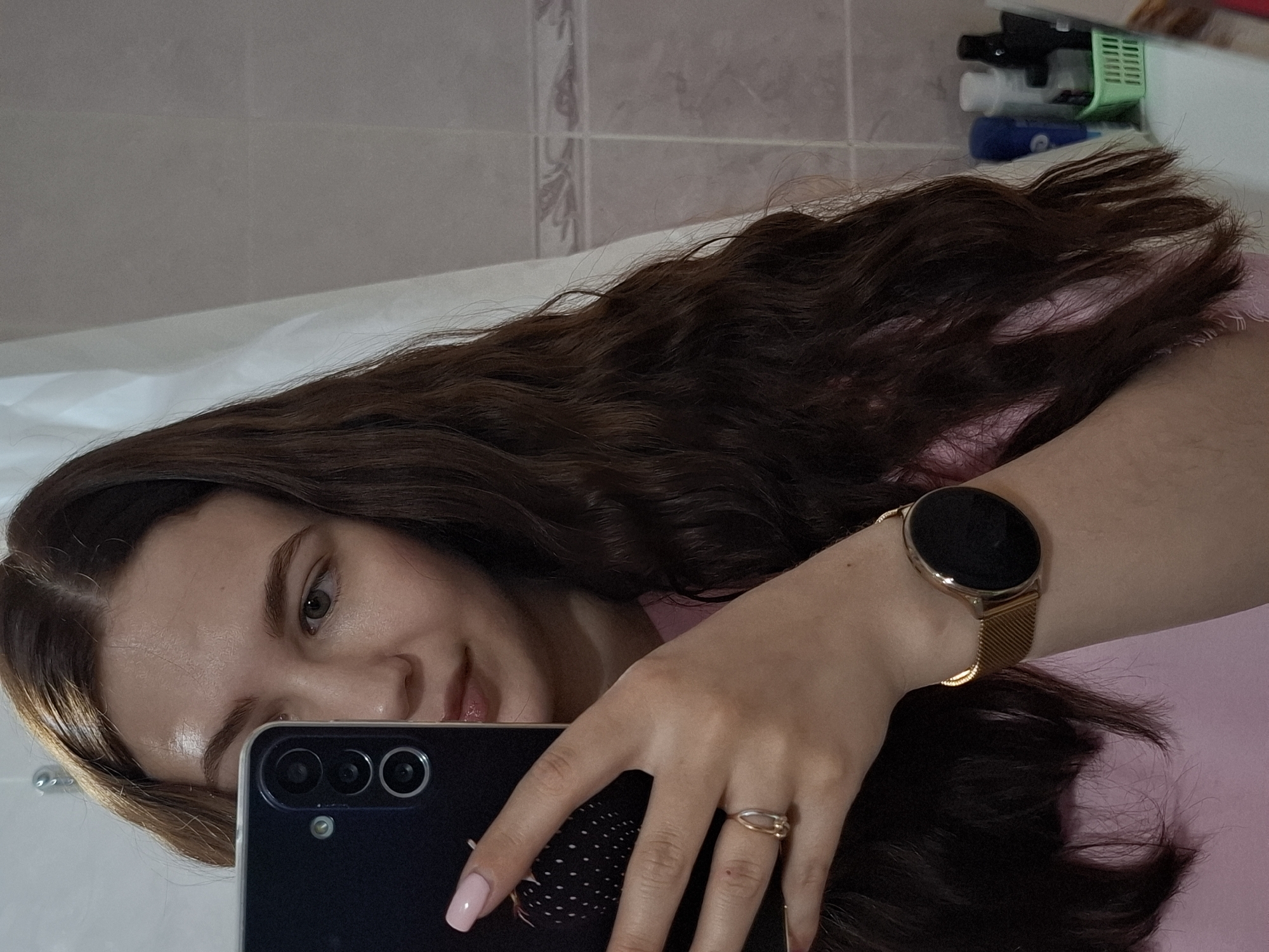 A young woman is depicted in a mirror selfie, taken indoors, likely within a bathroom in Chaikovsky, Russia. Her long, wavy, dark brown hair flows prominently across the frame, partially covering her pink top. She has a gentle, subtle smile and light-colored eyes, looking towards her reflection. Her forehead shows a slight glint, indicating soft lighting.
She holds a dark-colored smartphone in her left hand, which displays light pink nail polish on her fingers. The back of the phone features a triple camera module with a small flash. On her right wrist, she wears a gold-toned watch with a mesh band and a circular, dark face. A simple gold-colored ring adorns a finger on her right hand.
The background reveals light-colored, patterned wall tiles, characteristic of a bathroom setting. In the upper right corner, various toiletries in bottles are neatly arranged within a green caddy. A white surface, possibly the edge of a sink or bathtub, is visible in the foreground. No explicit text is visible in the image.