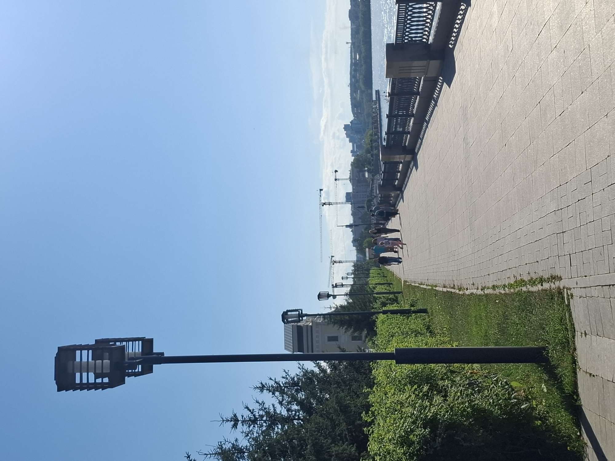This image depicts a sunny day along a paved waterfront promenade in Irkutsk, Russia. The sky is a clear, bright blue, suggesting pleasant weather. The promenade itself is paved with light-colored rectangular tiles, bordered by a grassy area with manicured bushes and trees.
Along the promenade, several modern street lamps with square, open-cage fixtures are visible, spaced out along the path. In the distance, a body of water, likely the Angara River, sparkles under the sun, with a decorative metal railing running along the edge of the promenade overlooking it. Beyond the river, the cityscape of Irkutsk is visible, including buildings and construction cranes, indicating ongoing development or modernization.
A few people are strolling along the promenade, enjoying the sunny day. They are dressed in casual summer clothing, and their presence suggests a leisurely activity. The overall atmosphere is peaceful and relaxed, characteristic of a pleasant day in a city park or recreational area. The time of day appears to be late morning or early afternoon, given the bright sunlight and the angle of the shadows. There is no visible text in the image.