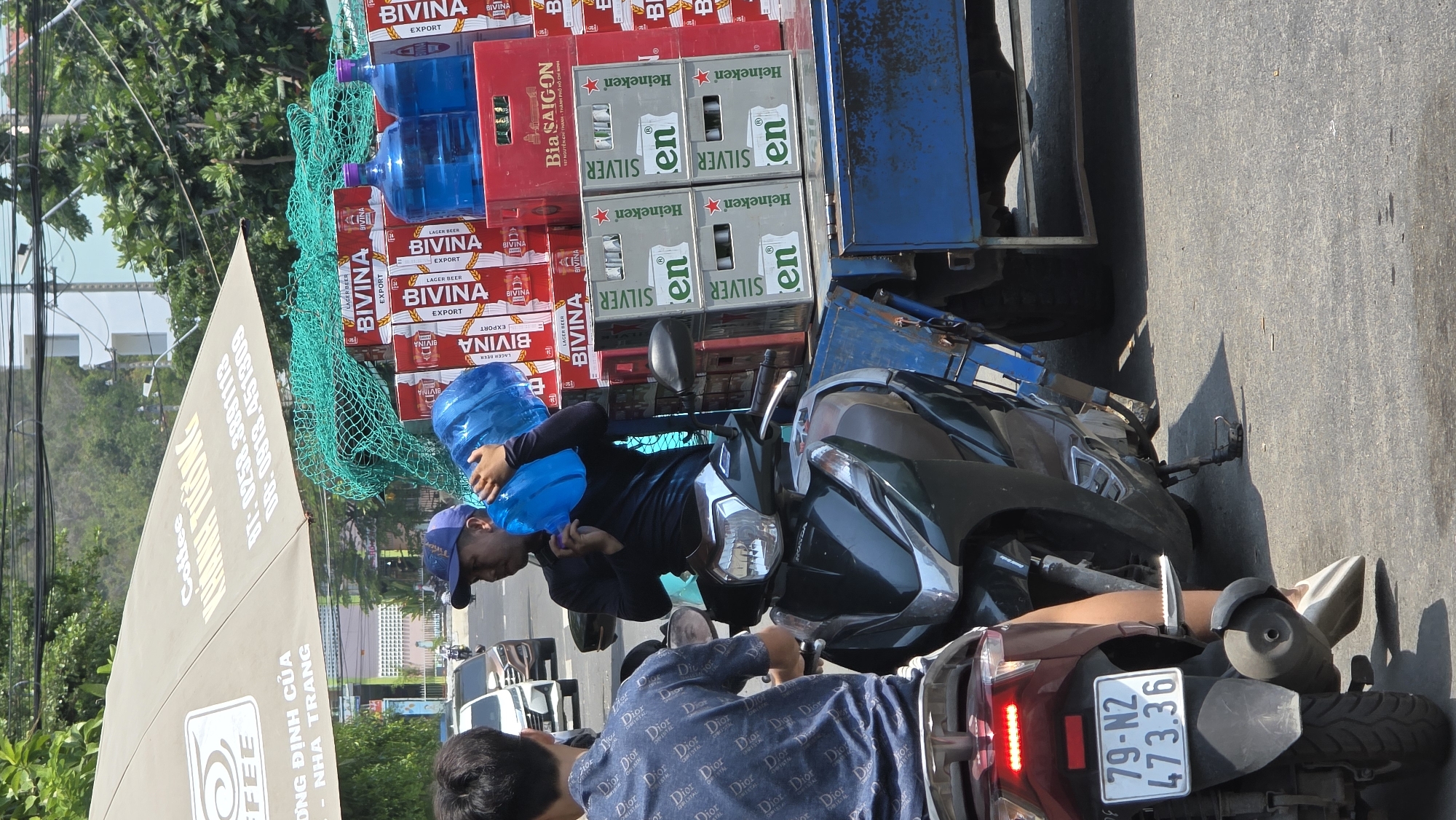 A partially rotated eye-level outdoor shot captures a street scene in Bac Nha Trang Ward, Vietnam. The primary focus is on a flatbed truck loaded with cases of beer and large water bottles, along with two adjacent motorbikes.
On the truck's bed are numerous cases of "BIVINA" beer, "Heineken" Silver beer, and red plastic cases labeled "Bia Saigon." Large blue water bottles are secured on top with a green mesh.
A person wearing a blue cap and a black shirt is positioned near the truck, holding a blue water bottle. In the foreground, a second person is on a motorbike, wearing a blue patterned shirt. The license plate of the motorbike reads "79-N2 473.36."
A sign with "Coffee KUNINH TUONG" in Vietnamese is on the left. The background shows a street, buildings, trees, and power lines. The scene appears to be captured during the daytime in bright sunlight.