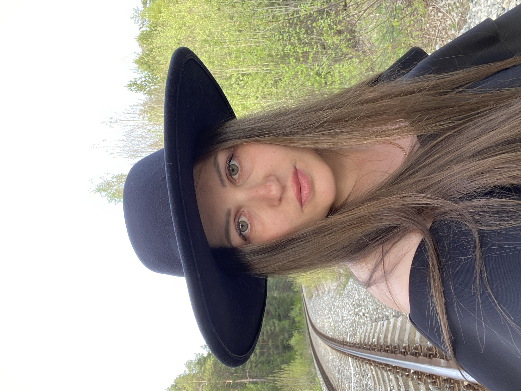 A young woman with long brown hair and a black hat is posing on a railway track. The tracks are flanked by greenery and trees, suggesting a location near the outskirts of Chaikovsky, Russia, possibly adjacent to a forested or less developed area. The woman is wearing a dark, possibly black, jacket or coat. She is looking towards the camera.