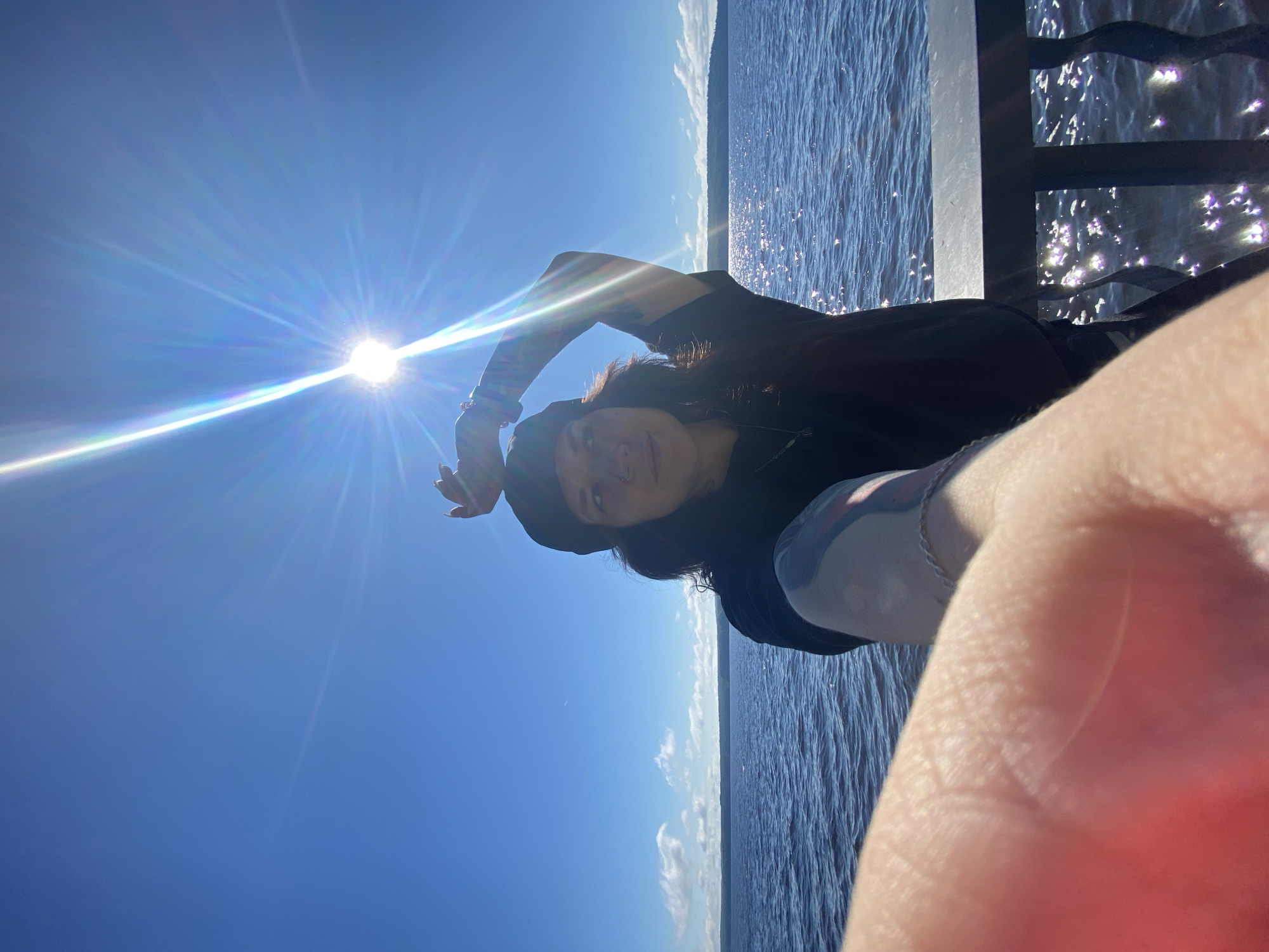 A young person with dark hair and a black beanie is taking a selfie outdoors. They are wearing a black t-shirt and have tattoos on their arms. The person is looking slightly to the right, with their left arm raised above their head. The background is a bright blue sky with the sun shining intensely, creating a starburst effect. Below the sky is a body of water, likely a river or lake, with gentle waves and sunlight reflecting off the surface. In the foreground, a person's hand is blurred, indicating it's holding the camera for the selfie. The scene appears to be on a pier or boardwalk, with a dark railing visible in the lower right corner. The overall atmosphere is sunny and bright. The location is identified as Chaikovsky, Russia.