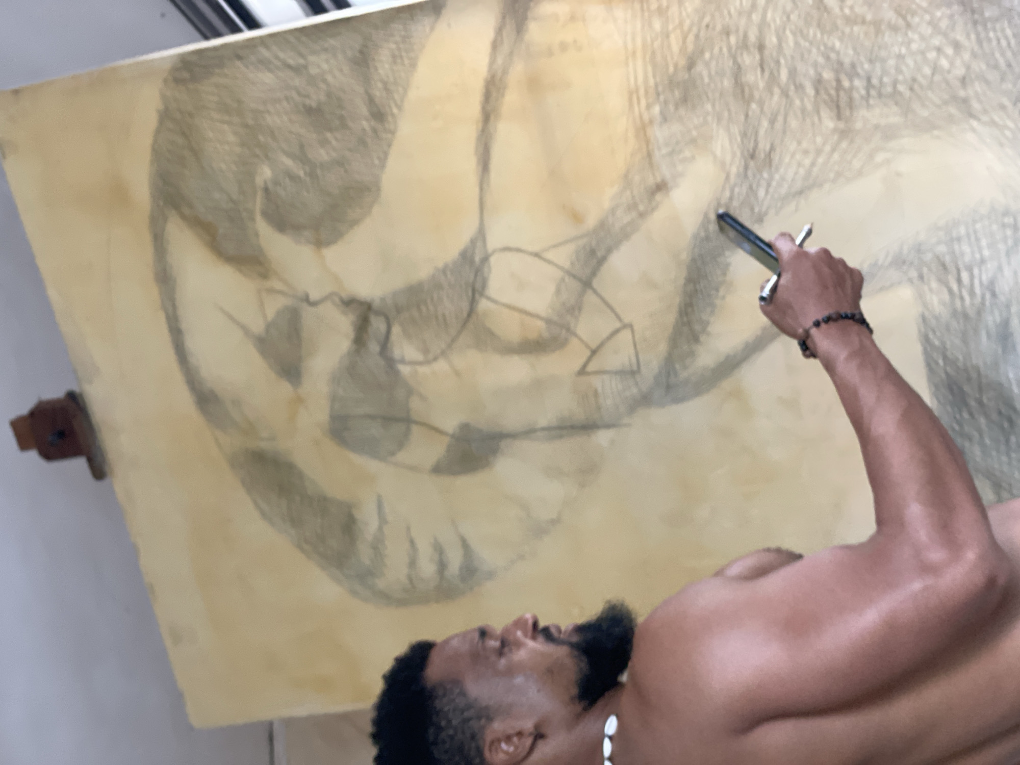 This vertical shot captures a male artist, likely in Ado, Nigeria, engrossed in drawing on a large sheet of paper or canvas. The man, with dark skin, a beard, and short dark hair, is shirtless, wearing a necklace made of white shells and a beaded bracelet on his right wrist. His muscular right arm is extended, holding a drawing tool (possibly charcoal or graphite in a holder) as he works on the artwork.
The drawing itself is abstract, featuring bold, sweeping lines, some geometric forms, and extensive cross-hatching in shades of gray and black against a light brown, possibly aged, background. It appears to be a large-scale piece mounted on an easel, though the easel structure is mostly obscured. The artist's gaze is directed towards his creation, indicating deep focus and concentration.
The setting appears to be an indoor studio or workspace, with a neutral-colored wall visible in the background. The lighting highlights the artist's skin, which has a slight sheen. There is no visible text in the image.