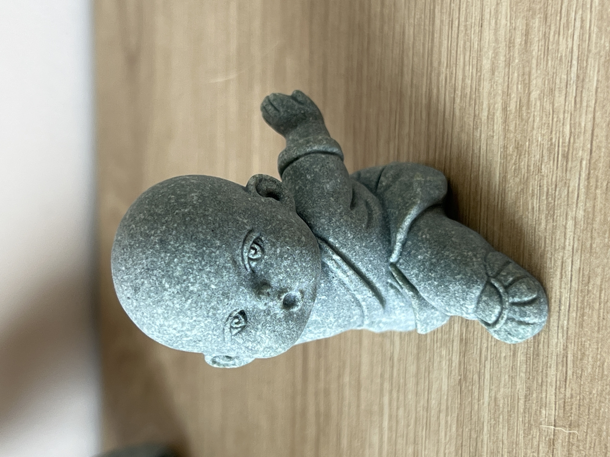 A gray, stone-like figurine of a baby lying on its side is centered in the frame. The figurine has a round head, detailed eyes, nose, and mouth, and a body clad in what appears to be a robe. The baby's limbs are slightly flexed. The figurine rests on a surface with a light brown wood grain pattern. The background includes a blurry, light beige or white vertical surface. The location, Nam Natural Heritage Area Trang, Vietnam, is not evident in this image.