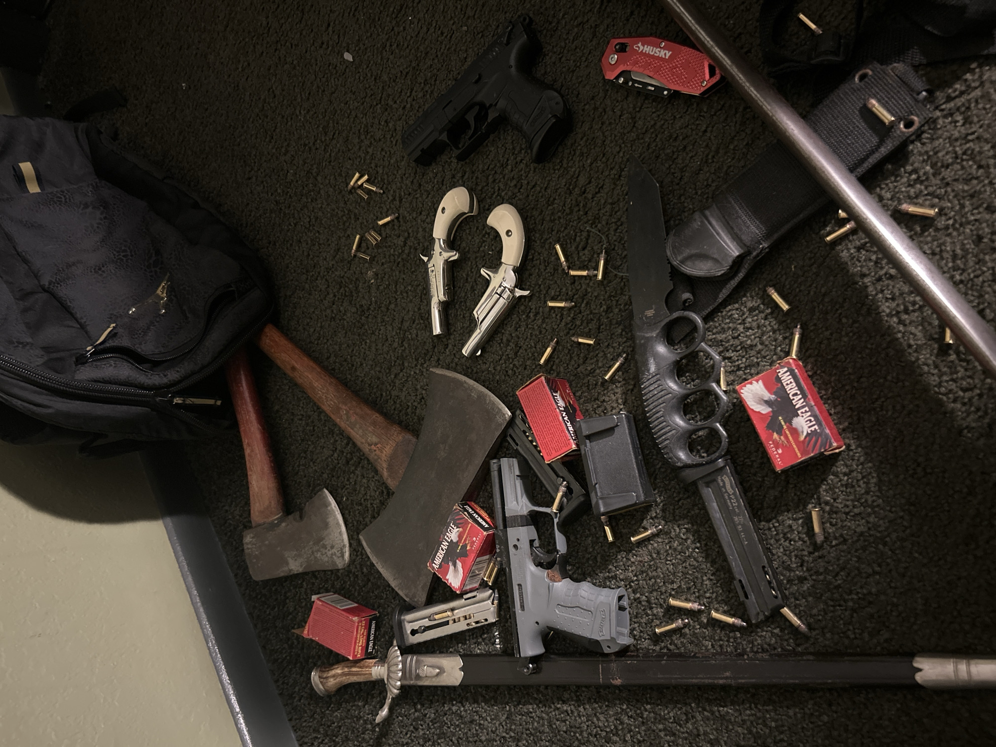 The image displays a collection of various weapons and related items laid out on a dark, textured surface, likely carpet. The scene does not depict any people or activity, but rather a static arrangement of objects.
Key items visible include multiple firearms: one black semi-automatic pistol, two silver derringer-style pistols with white grips, and one gray semi-automatic pistol with an inserted magazine. Scattered throughout the frame are numerous loose .22 LR ammunition cartridges. Several red boxes of "AMERICAN EAGLE" brand .22 LR ammunition, some with "FEDERAL" visible, are also present, along with additional magazines for the semi-automatic pistols, some appearing loaded.
Bladed weapons include two axes with wooden handles, a large black knife featuring a knuckle guard and a textured handle, and an ornate dagger or short sword with a decorative hilt and scabbard. Other items include a black backpack, a red "HUSKY" brand utility knife or multi-tool, and a metal pole or staff running diagonally across the right side of the frame.
The lighting suggests an indoor setting, possibly under artificial light. There are no specific cues within the image itself to determine the time of day or weather. The overall impression is that of a prepared cache or collection of various tools and weaponry. The location context provided is Cape Coral, United States of America.