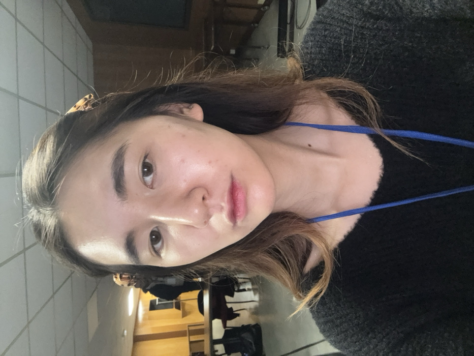 A woman with light skin, dark eyebrows, and brown eyes looks at the camera. Her lips are pink, and she has a mole on her neck. She wears a dark, possibly sweater-like, garment with a blue lanyard around her neck. Brown hair, partly tucked behind her ear with a leopard print clip, frames her face. The background is a room with tiled walls and wooden furniture, including chairs and a desk, visible in a slightly blurry perspective. The image is rotated 90 degrees.