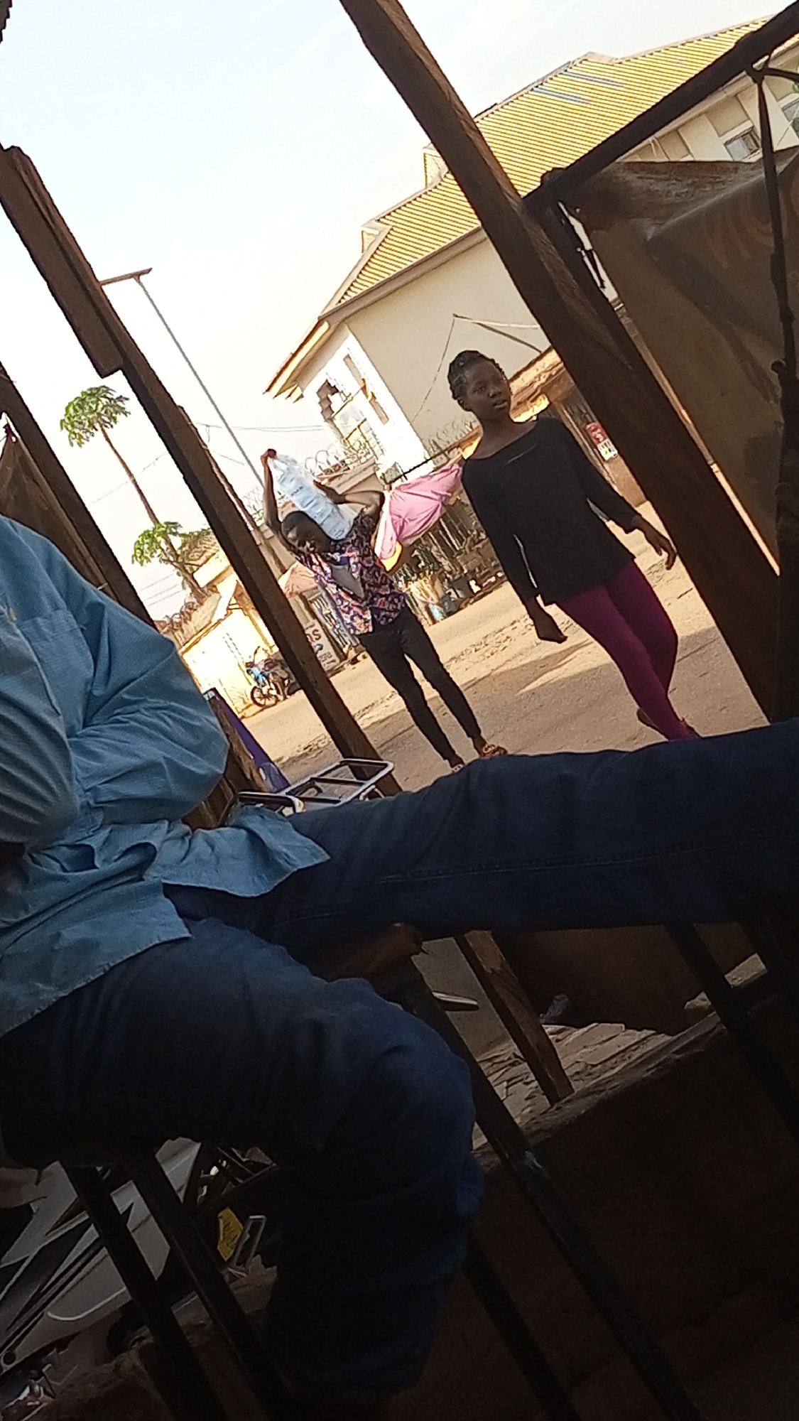The image, taken in what appears to be Sabon Tasha, Nigeria, shows a street scene viewed from an elevated, enclosed position. A person wearing a light blue shirt and blue jeans is partially visible in the foreground, occupying a seat. Beyond them, two people are walking on the street. One carries two large water bottles on their shoulders. The second person wears a black shirt and purple leggings. A motorcycle is seen parked on the street further away, and buildings are visible in the background. Street lighting and tree foliage are also present. The photograph's perspective is angled, as if taken from within a structure.