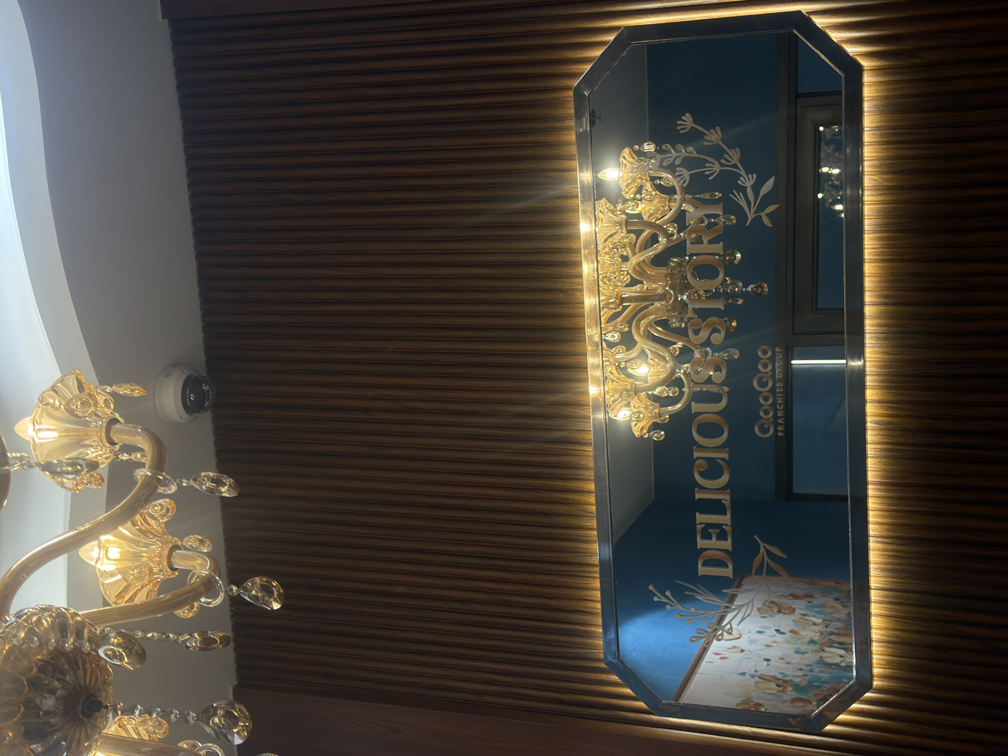 The image shows a vertically oriented, octagonal mirror hanging on a wall with horizontally oriented wood panels. The mirror has a blue background with a gold floral design on its upper portion and the words "DELICIOUS STORY QOOQO FRANCHISE GROUP" in gold lettering. Inside the mirror's reflection is a glimpse of the room it is in, including a picture with blue and white tones.
To the left of the mirror is a chandelier with golden fixtures and transparent crystals. On the wall between the chandelier and the wood panels, there is a round, white security camera. The overall lighting in the scene is warm, creating a cozy and luxurious feel.
The location is Nha Trang Ward, Vietnam.