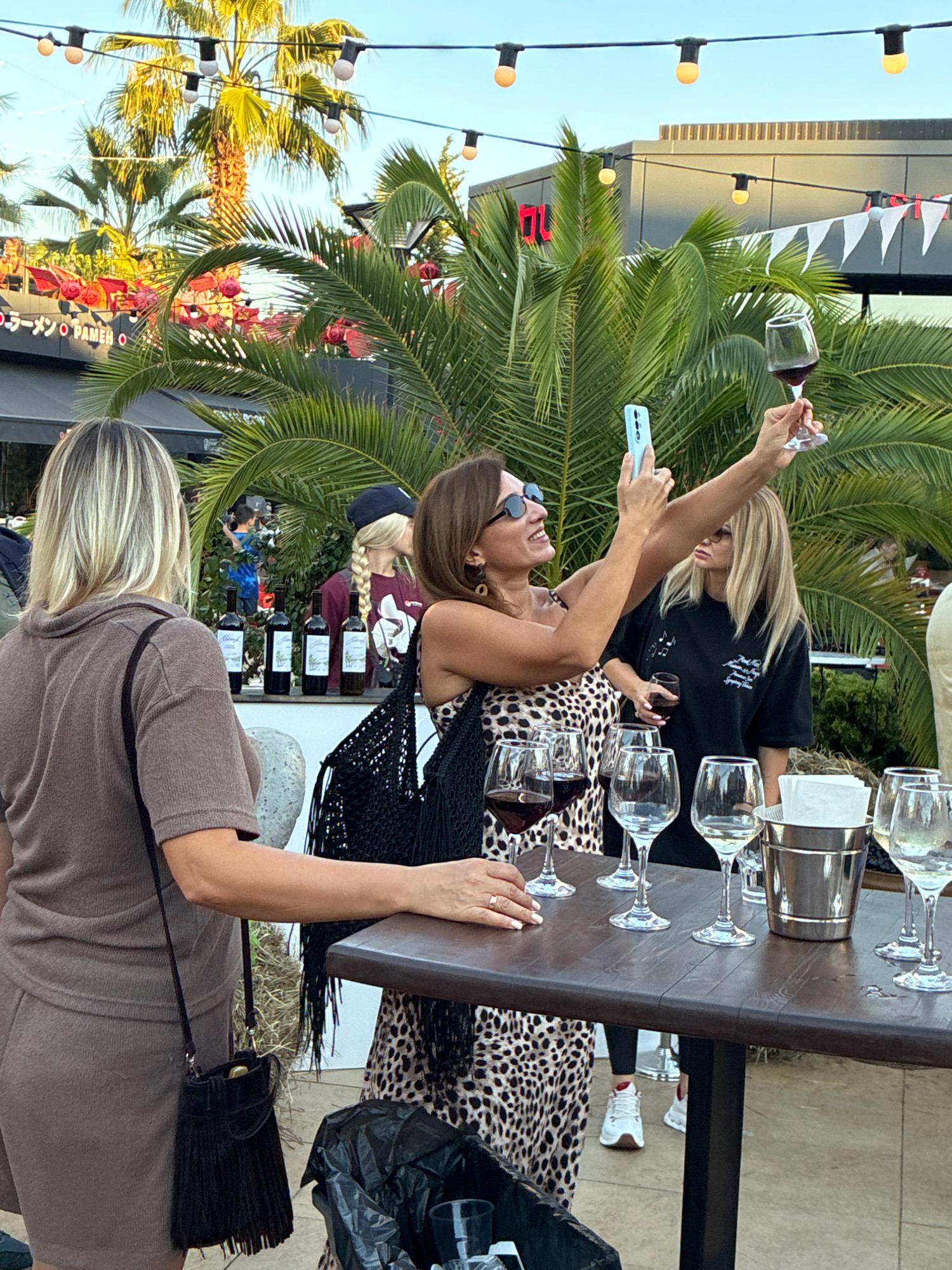 In an outdoor setting with palm trees and string lights, three women are gathered around a table laden with wine glasses. The woman on the left, wearing a brown outfit and carrying a fringed purse, stands with her back to the camera. A woman in the center, dressed in a leopard-print dress, holds up a phone to take a selfie with a glass of red wine. To her right, another woman in a black t-shirt also holds a glass of wine. Behind them, a display of wine bottles is visible. The ground is paved, and a trash can is present in the foreground. The scene appears to be at a wine tasting event, possibly in the city of Sochi, Russia, given the palm trees and general ambiance.