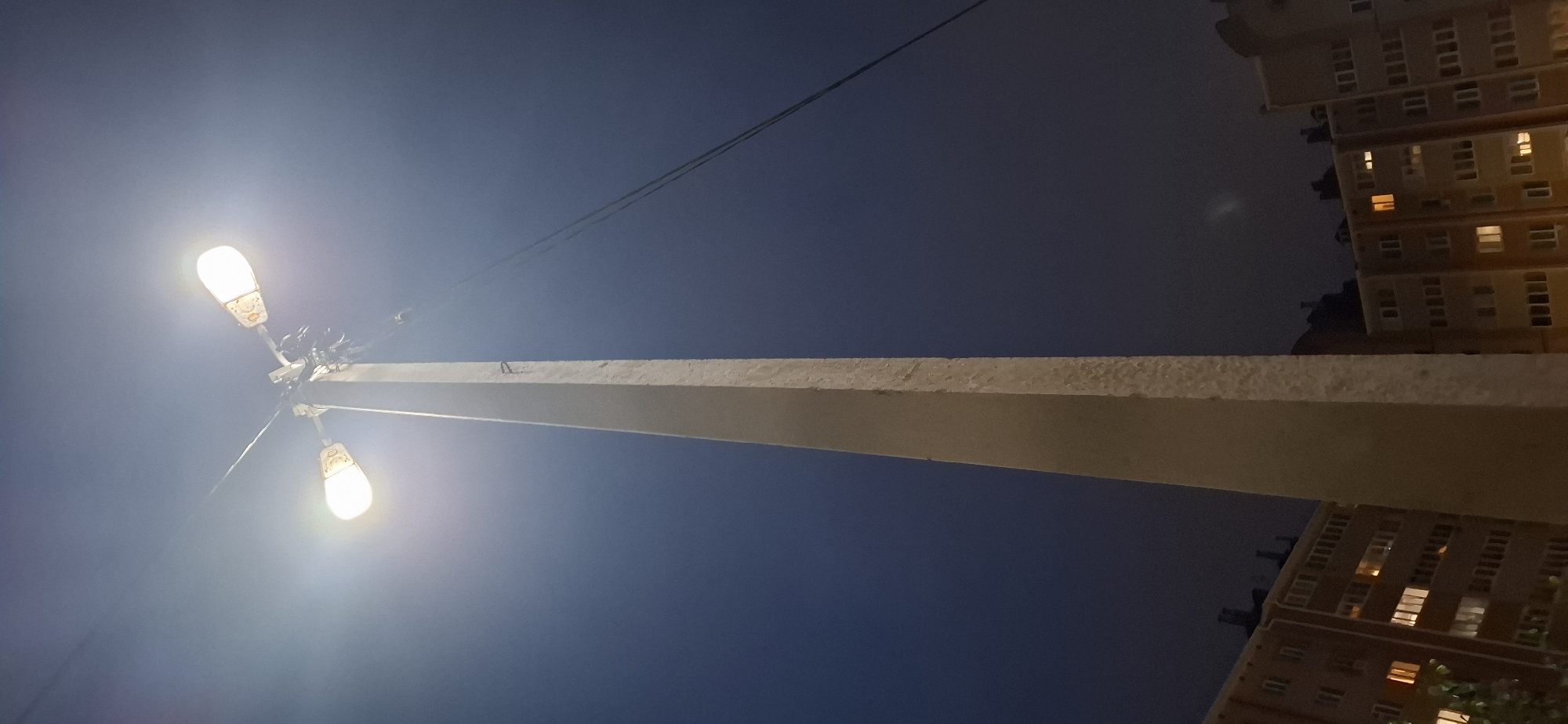 The image shows a tall, concrete lamppost with two illuminated lamps at its top, set against a dark blue, night sky. The lamppost is positioned in the foreground, dominating the center of the frame. On either side of the lamppost, the tops of apartment buildings are visible, with several windows lit from within. The environment appears to be urban, likely in Medvedevo Urban Settlement, Russia, given the architectural style of the buildings. No people or specific actions are depicted in the scene. The lighting suggests it is nighttime.