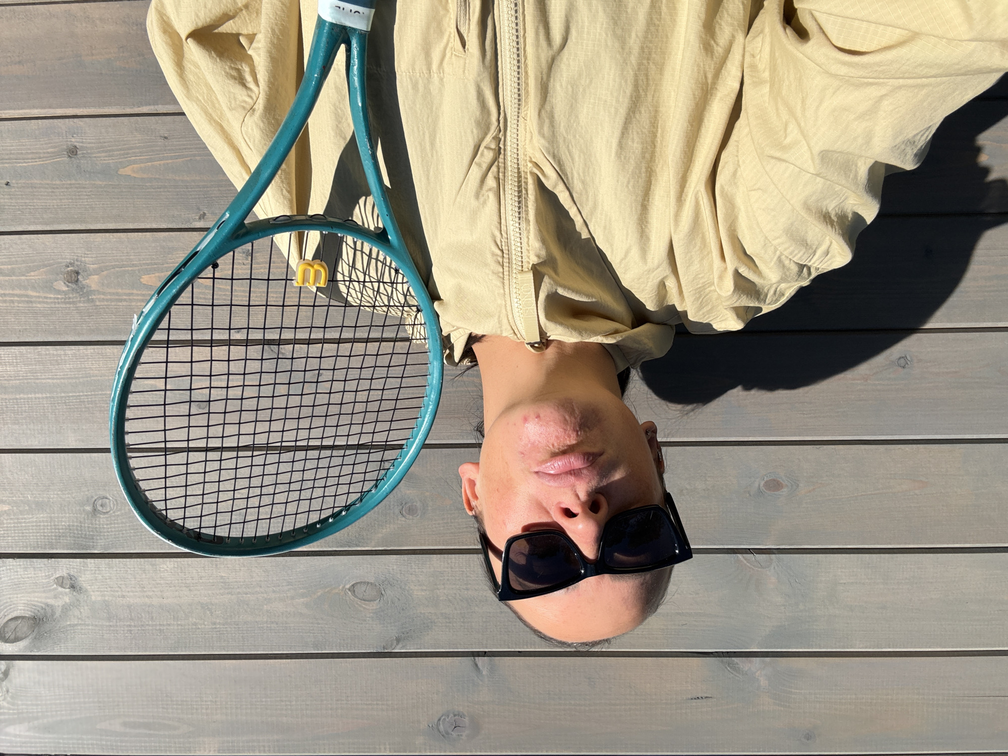 A woman wearing sunglasses and a light-colored hooded jacket holds a teal tennis racket in front of a weathered wooden fence. The woman is looking upwards with her lips puckered. A shadow of the woman and the tennis racket is cast on the fence to her left. The scene appears to be outdoors on a sunny day. The location is Chaikovsky, Russia.
