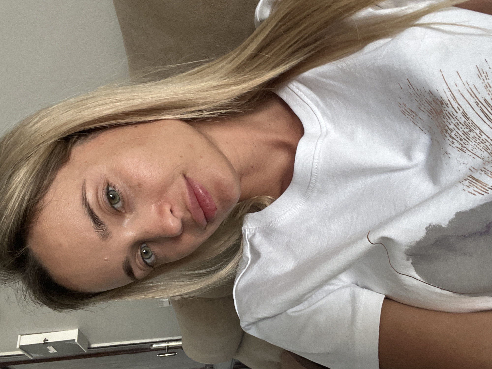 A close-up selfie of a woman with blonde hair and green eyes, wearing a white t-shirt. She is seated on a light brown couch in an indoor setting, possibly a hotel room or apartment. The background shows a grey wall and a part of a closet or cabinet. The lighting suggests it is daytime. There is a stain on her t-shirt. The scene is not specifically identifiable as Sharm El Sheikh, Egypt, or any other city.