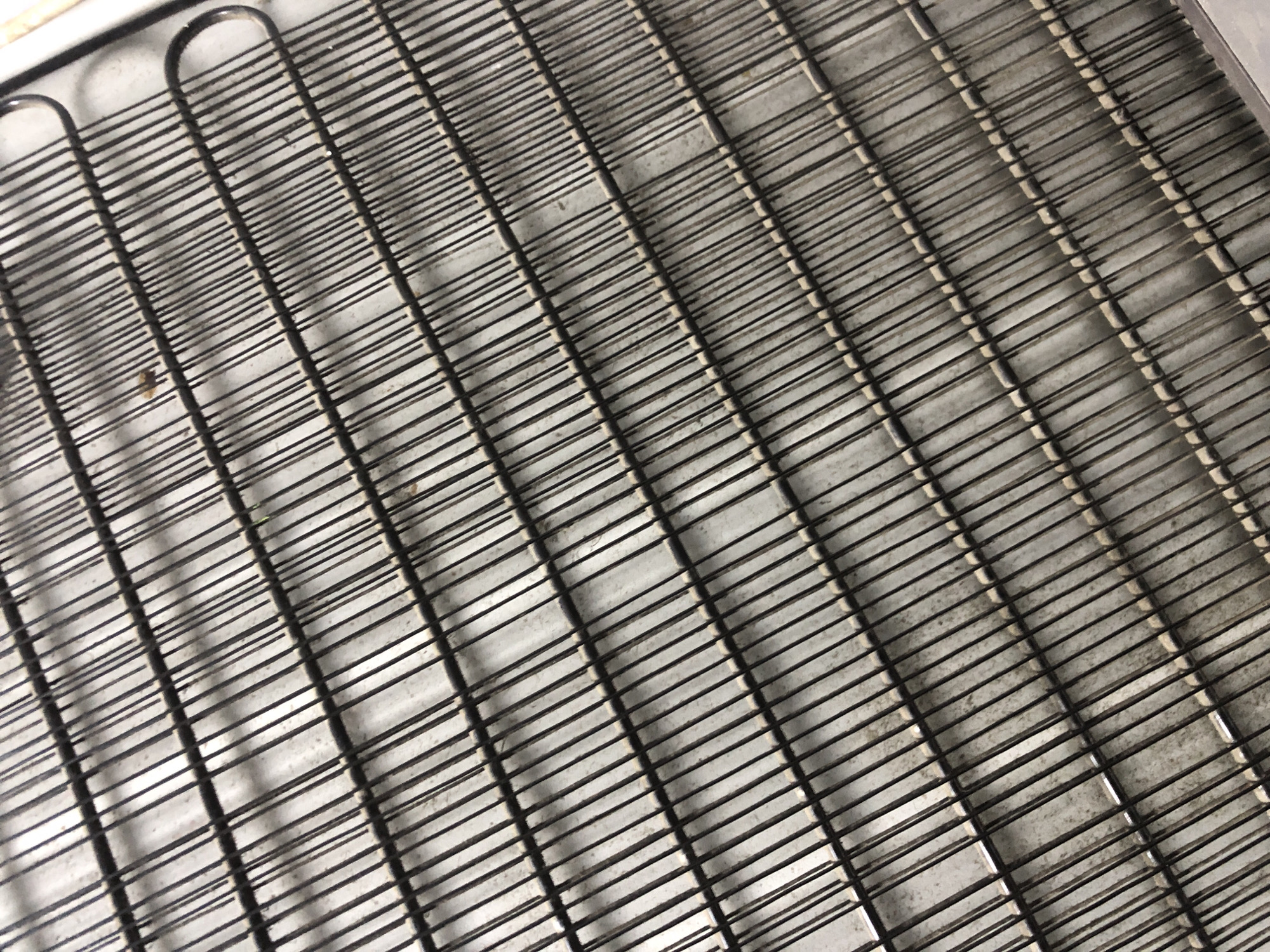 The image shows a close-up, high-angle view of a refrigerator condenser coil. The coil is made of thin, dark metal tubes arranged in a parallel, serpentine pattern against a light gray background that appears to be a wall or floor. The tubes are spaced closely together and are slightly dusty. There are a few small, dark specks, possibly debris or insect fragments, visible on some of the tubes. The overall impression is one of utilitarian mechanical components. The location in Ado, Nigeria is not ascertainable from the image alone.