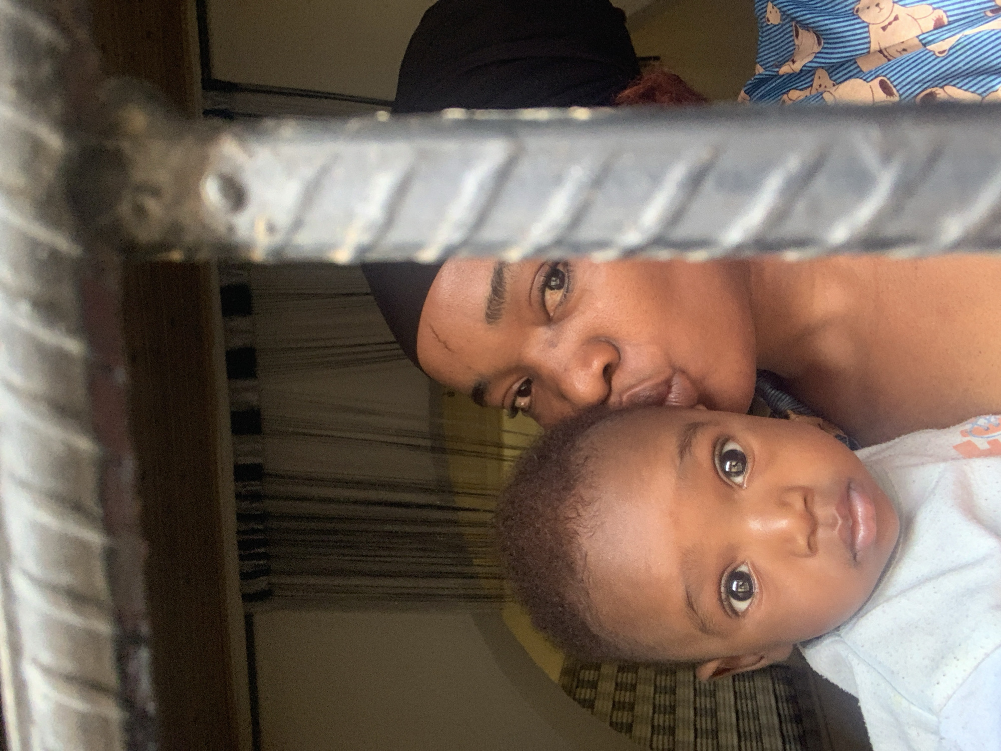 A portrait image, presented in a rotated orientation, captures an intimate moment between an adult female and an infant indoors. A thick, dark metal bar runs diagonally across the foreground, partially obscuring the view of the subjects.
The adult, visible towards the top-left of the frame, has dark skin and wears a black head covering. She is looking towards the camera while gently leaning in, seemingly to kiss the infant’s head or cheek. A portion of her blue and white striped garment, adorned with teddy bear motifs, is visible in the background.
The infant, positioned towards the bottom-right, has dark skin, short dark hair, and large, dark, expressive eyes, looking directly at the viewer. The infant is dressed in a light-colored top. In the background, behind the infant, is a pattern of horizontal stripes, likely from a curtain or blind. The overall scene conveys a tender bond between the two individuals, possibly a mother and child, within an indoor setting in Barakin Akawo, Nigeria. There is no visible text.
