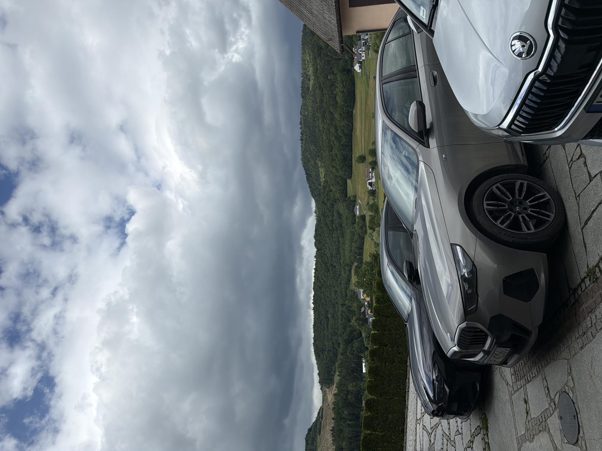 The image displays a frontal view of three cars parked on a paved surface. The leftmost car is black, followed by a silver car, and the rightmost car is also silver. In the background, a dense green forest covers rolling hills under a cloudy sky. Several houses are visible scattered among the hills. A tall, manicured hedge runs along the left side of the frame. The car on the left, a black BMW, is partially obscured by the silver car in the middle. The silver car in the middle is a BMW sedan, with its front grille and headlights clearly visible. The silver car on the right, a Skoda, shows its grille and hood. The license plate on the middle BMW reads "OP 4206U". The location is identified as Męcina, Poland.